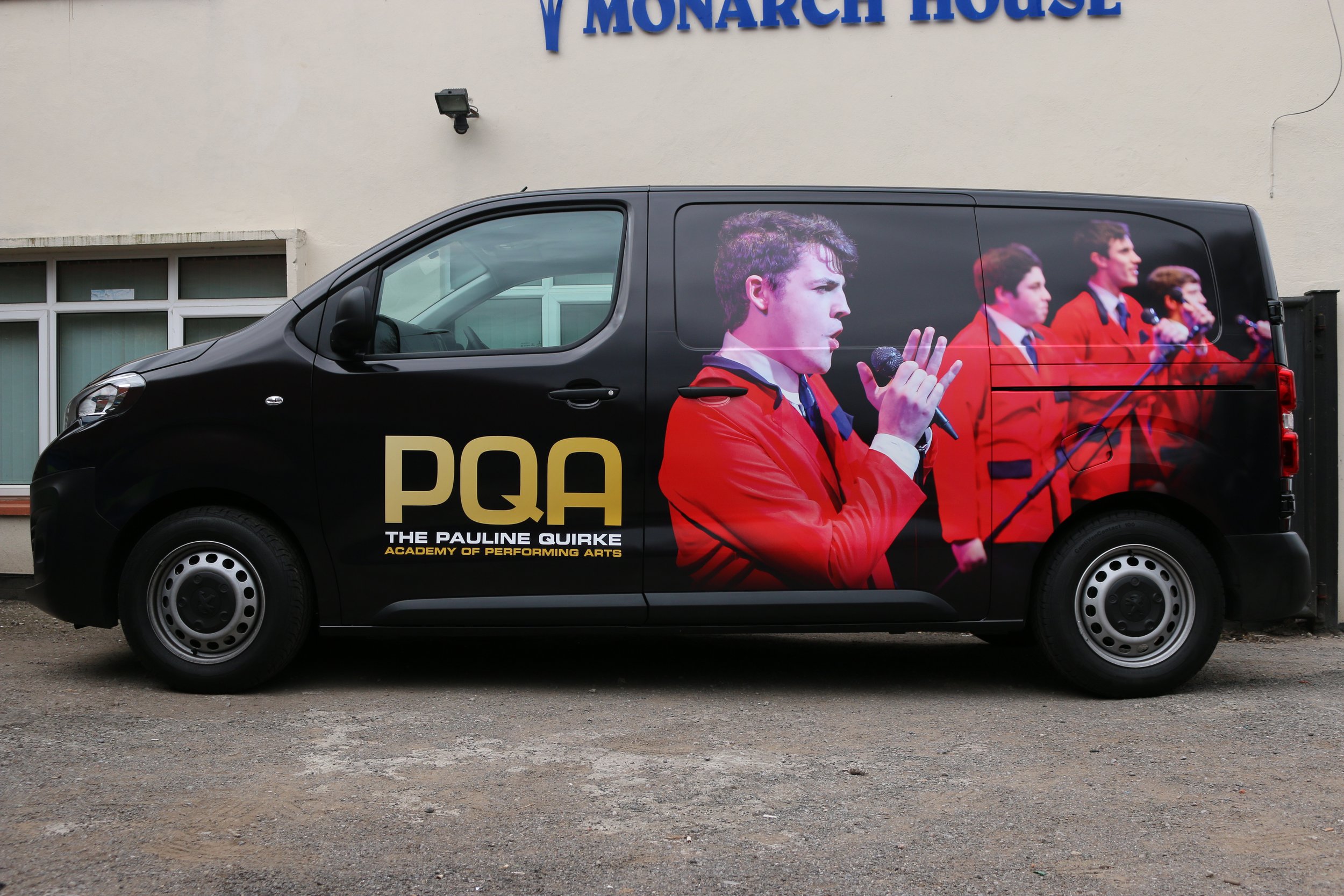 Black van with a large poster of a woman singing, wearing a red blazer, holding a microphone, on its side. The van is parked in front of a beige Building with a sign that reads "Monarch House" in blue letters.