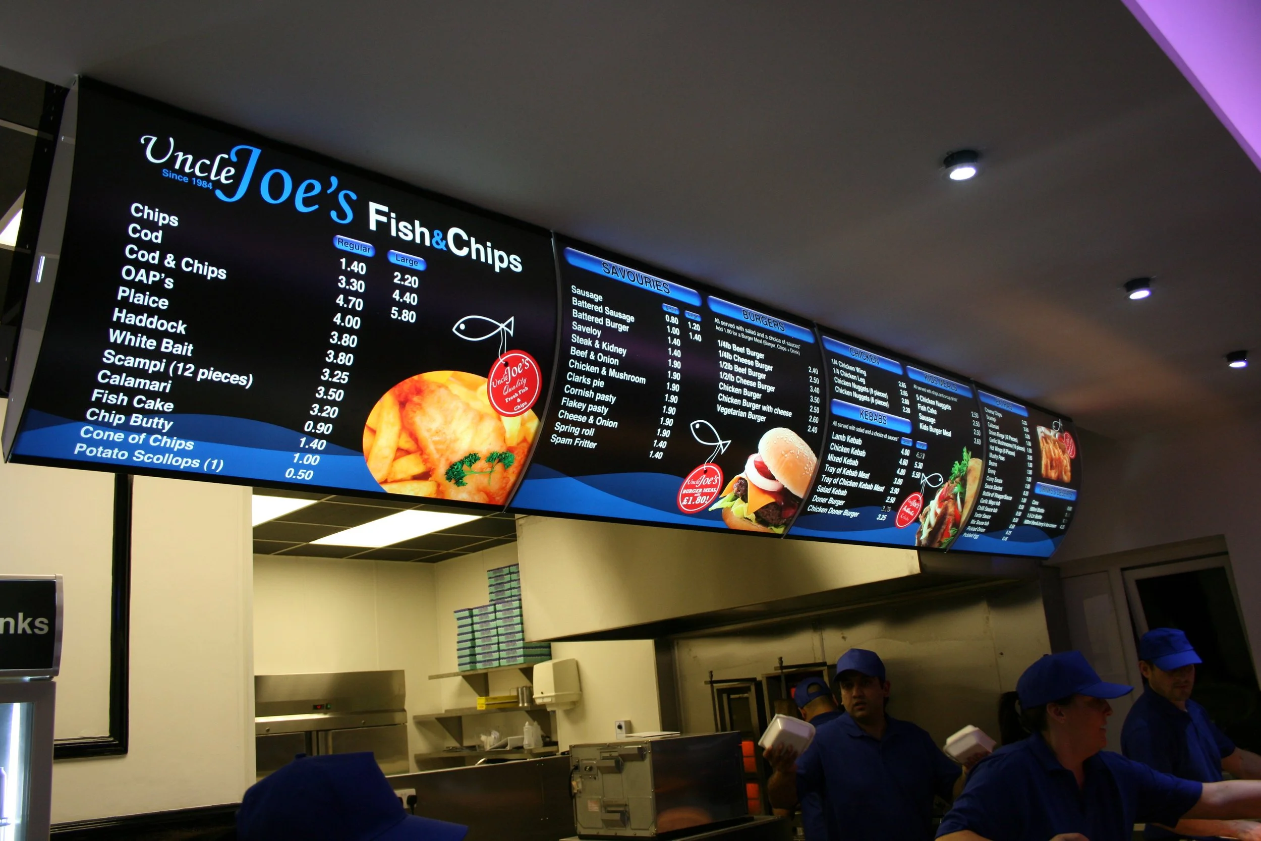Digital menu board at Uncle Joe's Fish and Chips restaurant showing food prices and items, with staff wearing blue uniforms and caps below.
