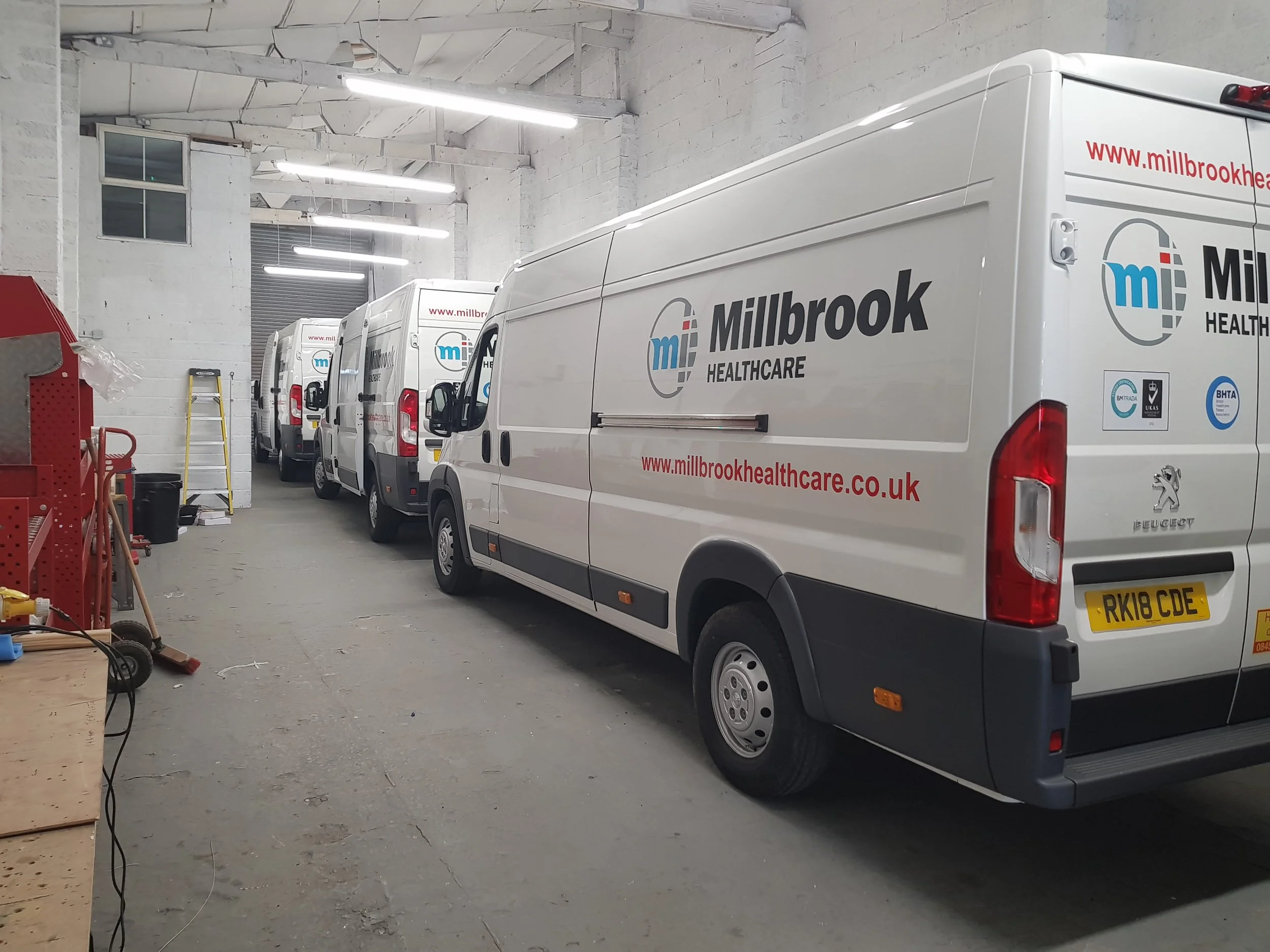 Interior of a warehouse or garage with white walls, several white delivery vans parked inside, some with company branding for Millbrook Healthcare. Fluorescent lights hang from the ceiling, and there are various tools and equipment along the left sid