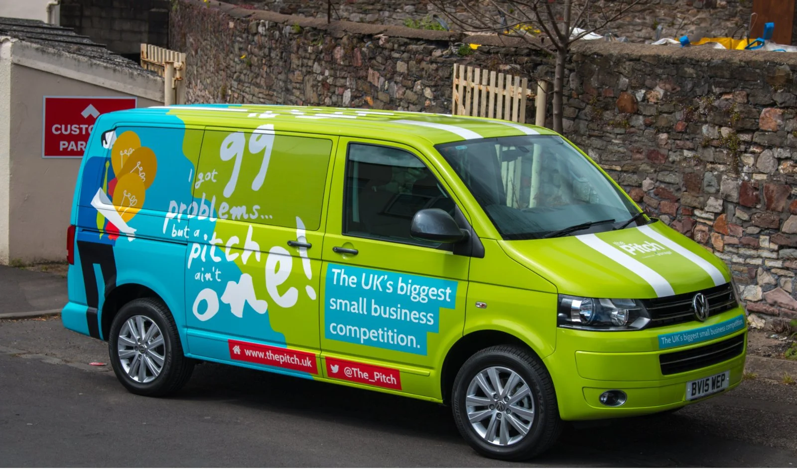 Green and blue van with promotional graphics for The Pitch, claiming it is the UK's biggest small business competition, parked beside a stone wall.