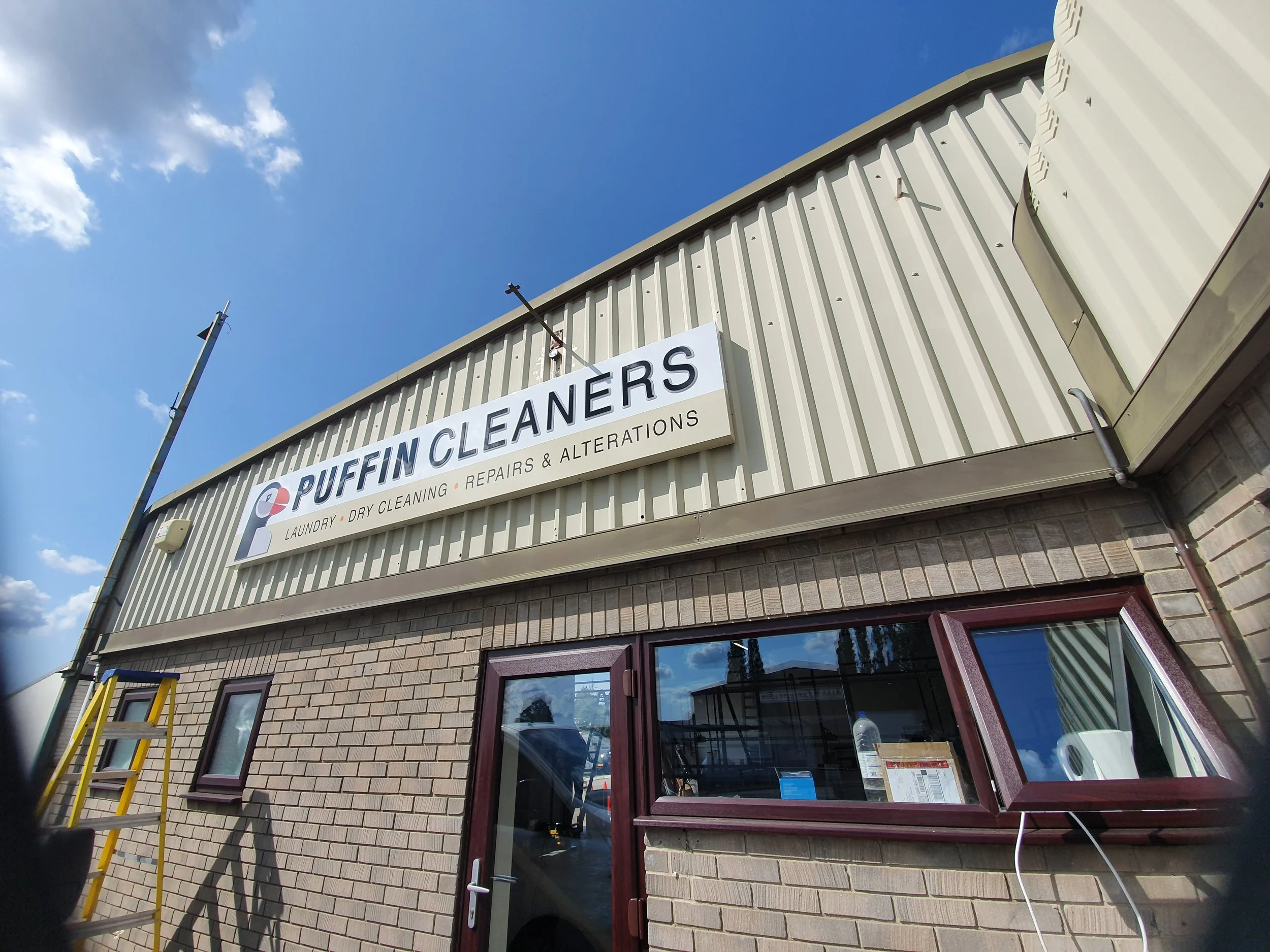 Exterior view of a commercial building with a sign that reads 'PUFFIN CLEANERS'. The building has a beige brick facade, a window with a burgundy-colored frame, and a door. A ladder is leaning against the wall near the window. The sky above is blue wi
