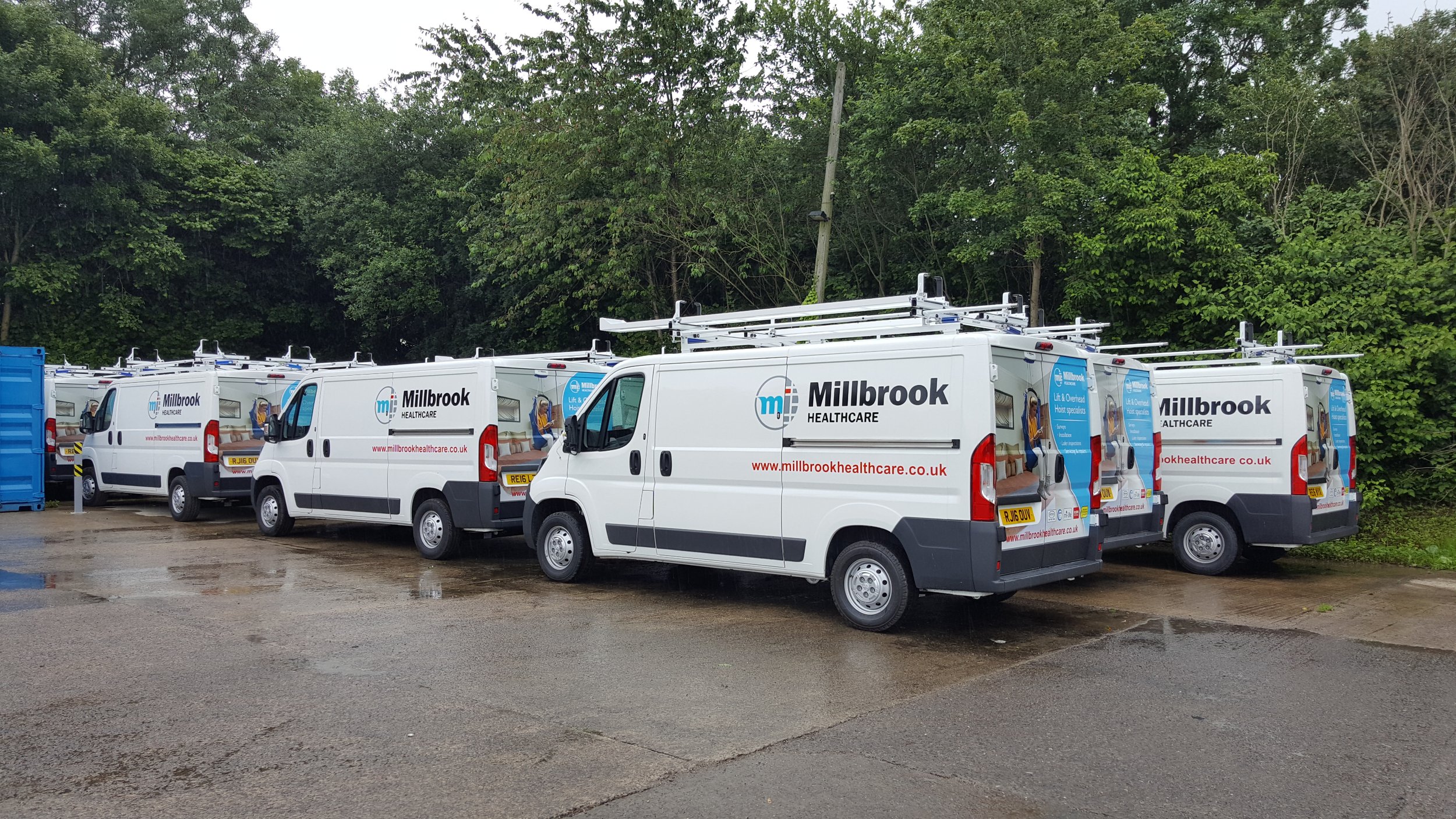 Several white healthcare vans with Millbrook Healthcare branding parked on a wet, muddy surface near a green woodland area.