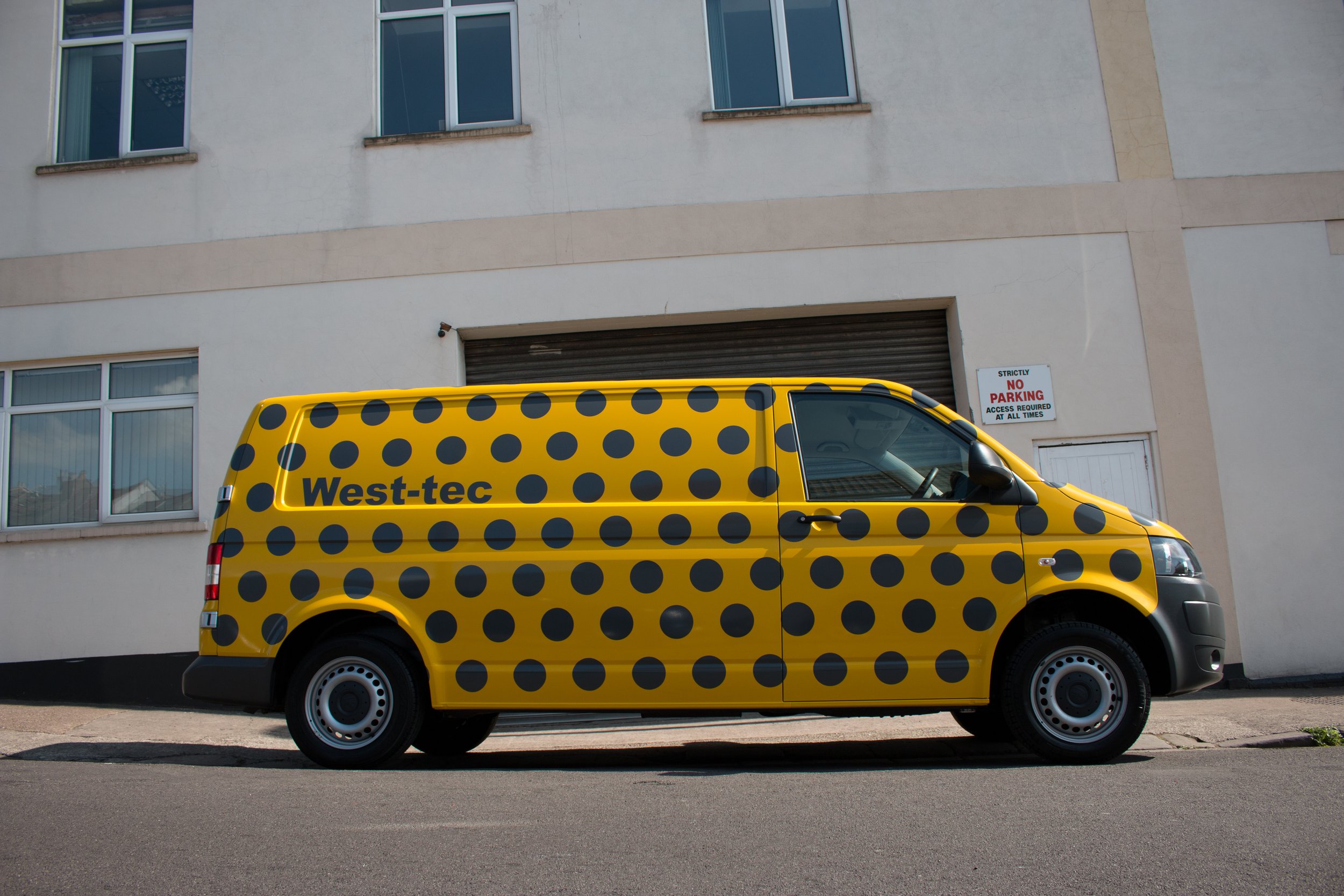 A yellow delivery van with black polka dots parked on the sidewalk in front of a beige building with multiple windows. A sign on the building states 'Strictly No Parking' and indicates access is required at all times.