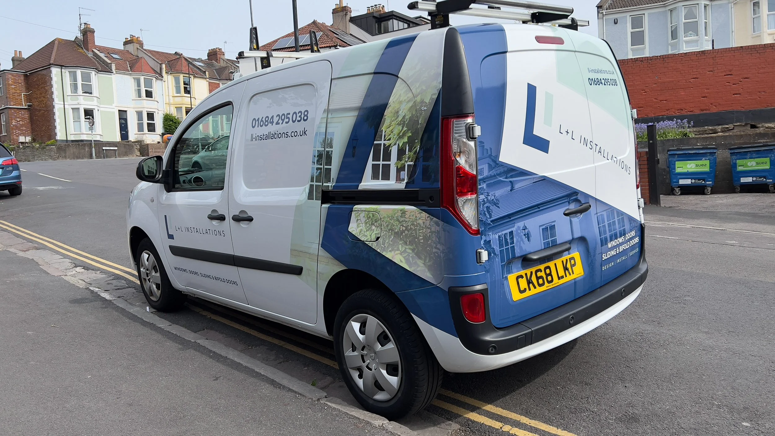 A white commercial van parked on the side of a street with colorful residential buildings in the background. The van has branding for L+L Installations, including a website and phone number, and features images of windows and doors.