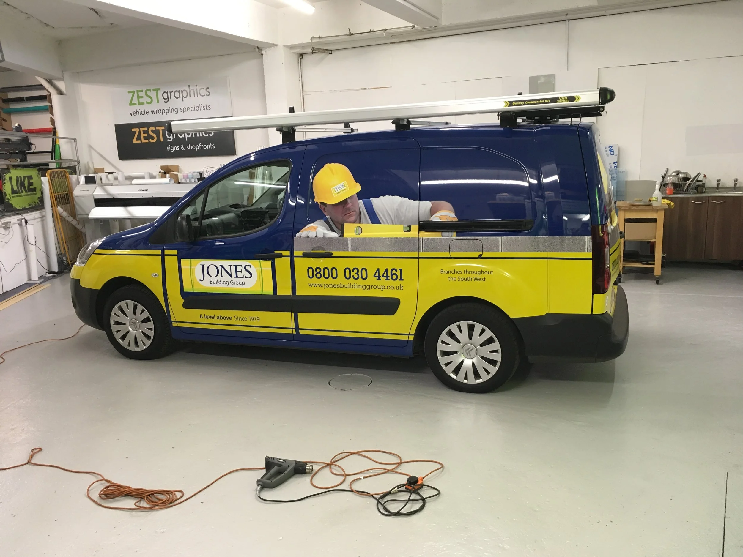 A worker wearing a yellow safety helmet and white shirt is leaning out of a blue work van with yellow and gray stripes, parked indoors. The van has the logo and contact details of Jones Building Group. The workplace has various tools, equipment, and 