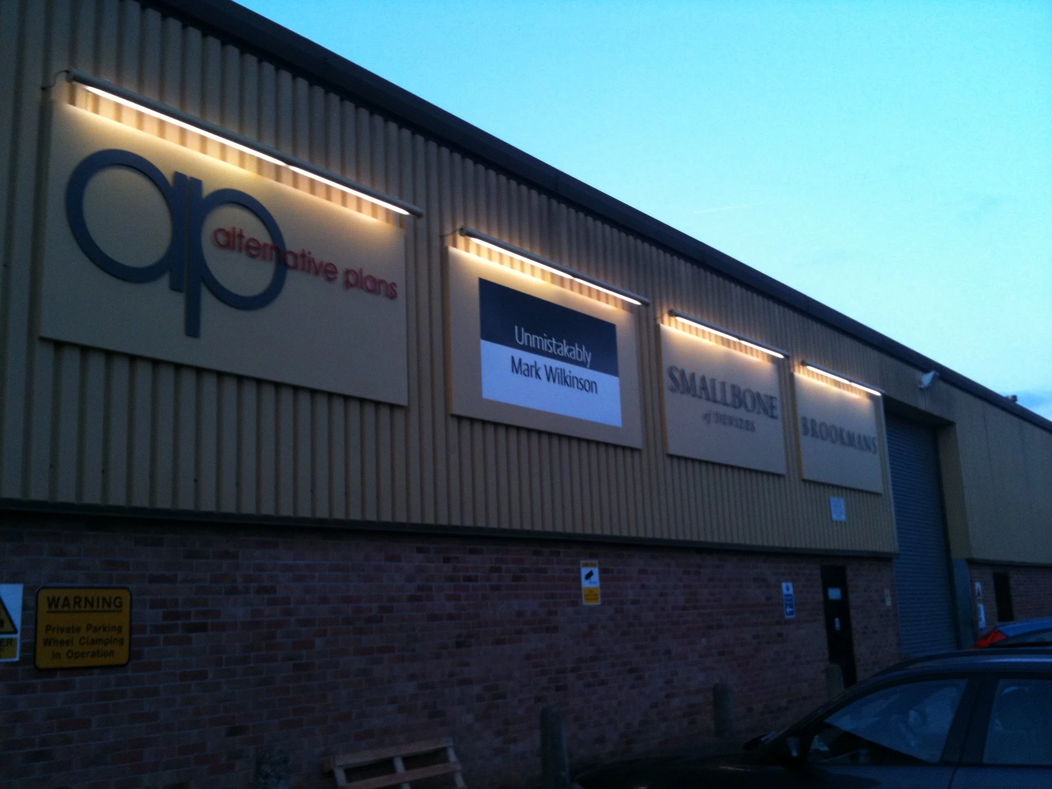 The exterior of a retail store during dusk or dawn, with illuminated signs for 'alter native plans,' 'Unmistakably Mark Wilkinson,' and 'Smallbone of Devizes,' and a yellow parking warning sign at the bottom.