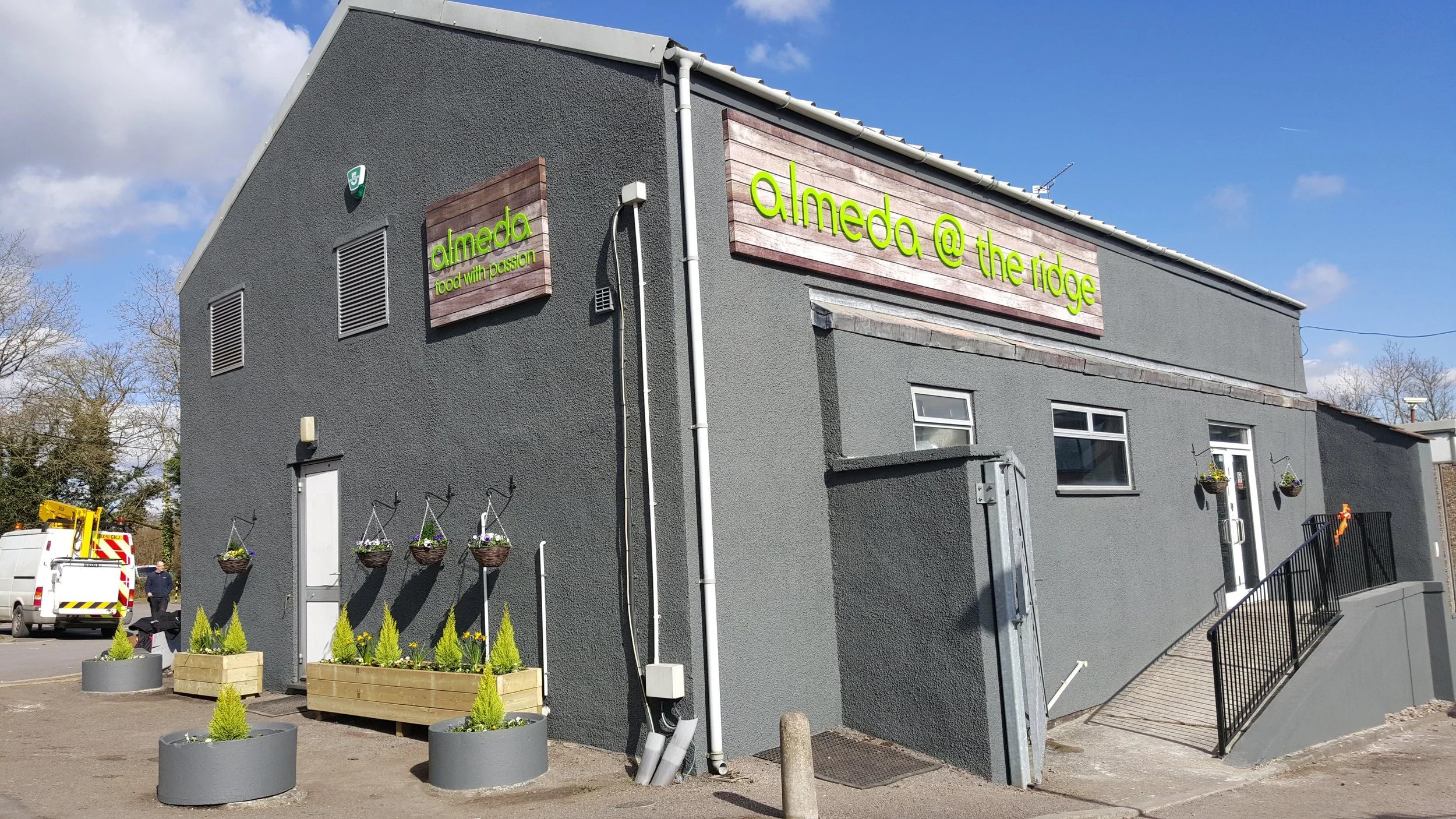 A gray building with a wooden sign that reads 'almeda @ the ridge' and 'food with passion' in green letters. The building has small windows, flower baskets, and planters with small greenery, with a parking lot and a person in the background.