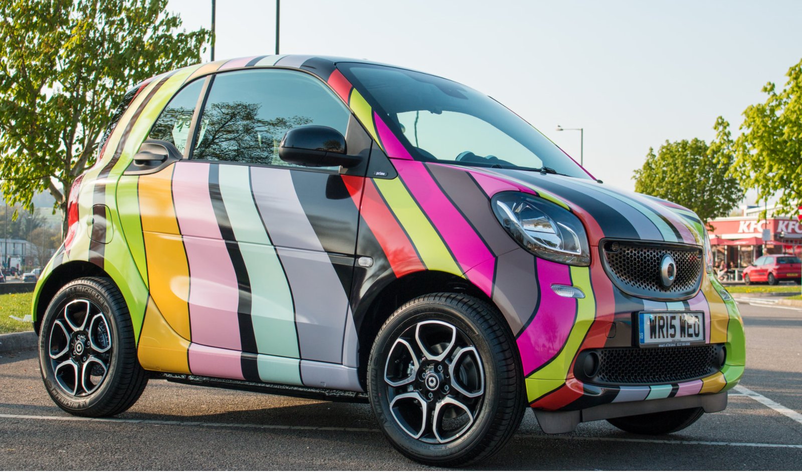 A small compact car with a colorful, striped wrap parked in a parking lot with trees and a fast food restaurant in the background.