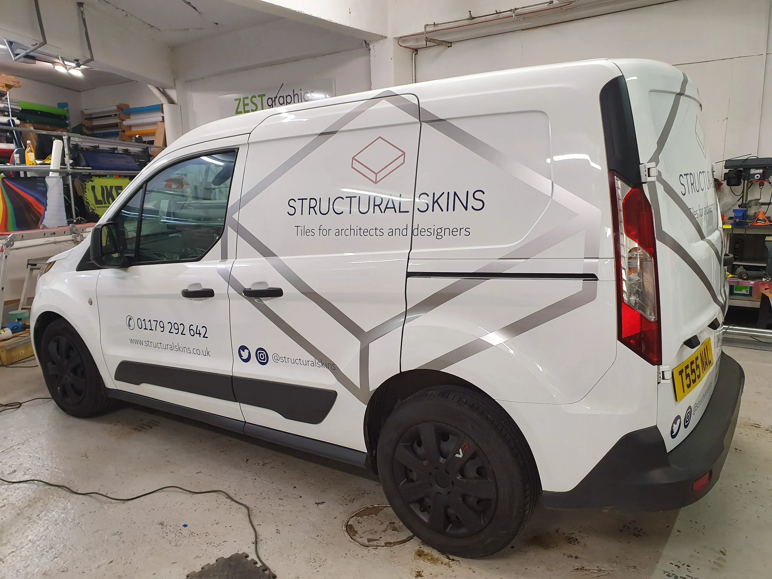 White commercial van with branding for Structural Skins, a company selling tiles for architects and designers, parked inside a workshop or storage area. The van's branding includes a logo of a cube and social media icons.