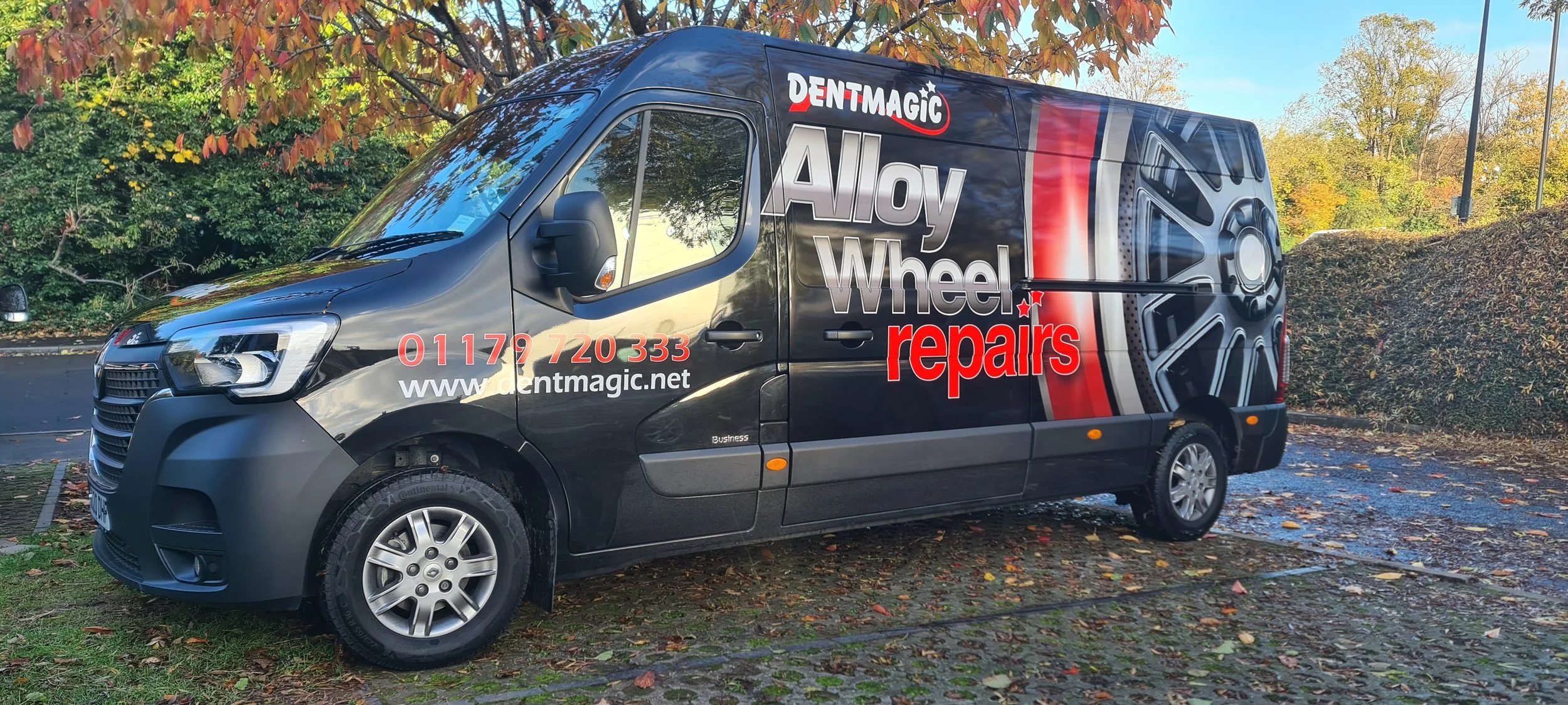 A black van with advertisement for alloy wheel repairs, featuring a large image of a wheel on the side, contact number, website, and company logo, parked on a wet pavement with autumn trees in the background.