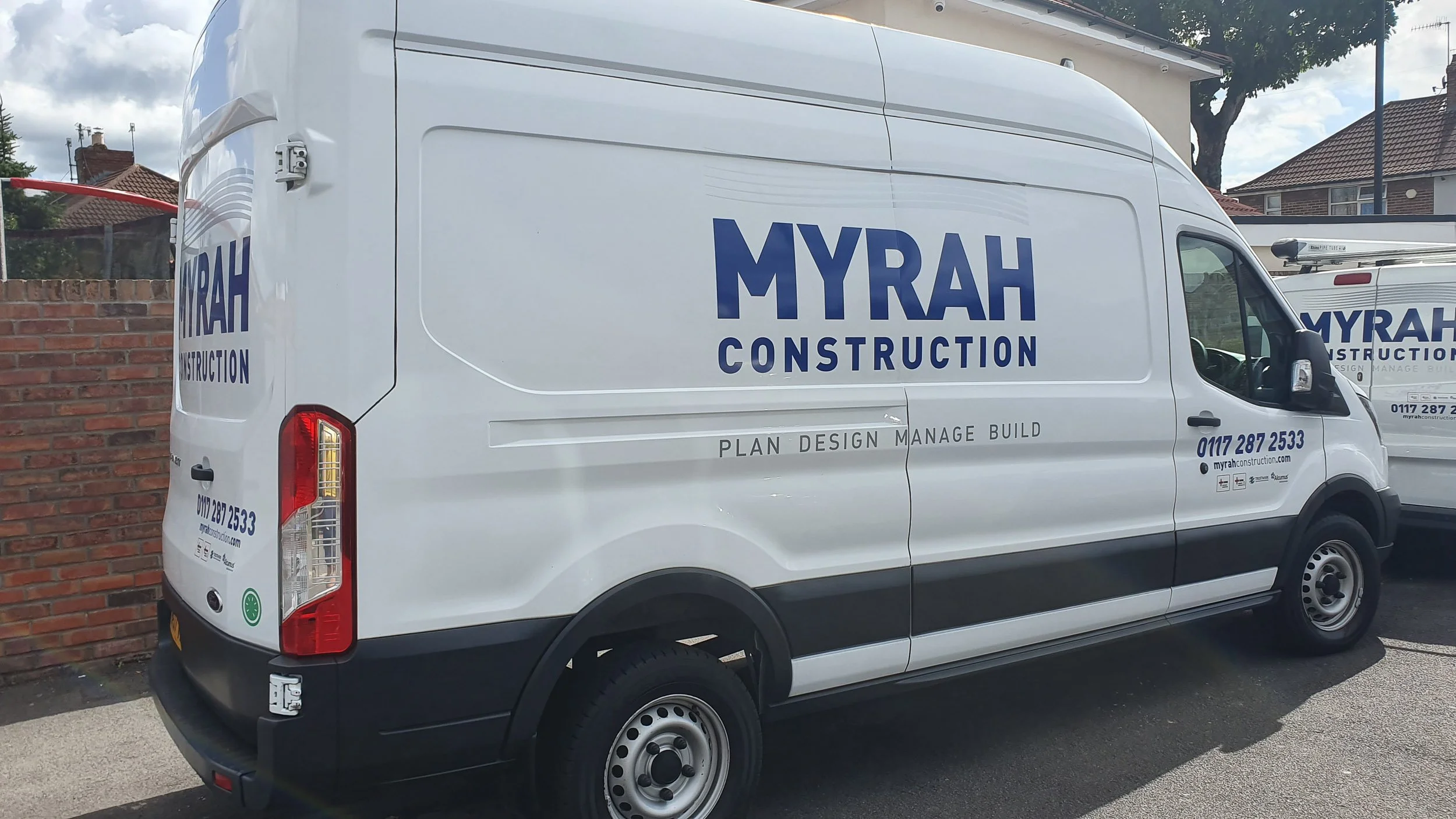 White construction service van parked on a street with the company name 'MYRAH CONSTRUCTION' in blue letters on the side. The van displays services like 'PLAN DESIGN MANAGE BUILD' and contact information, with residential houses and trees in the back