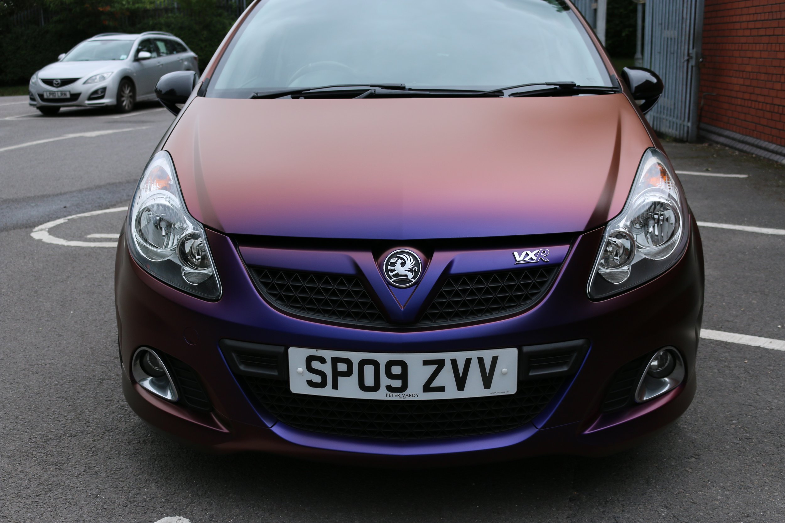 Front view of a maroon Vauxhall hatchback car parked in a parking lot, with a silver car in the background and a brick building on the side.
