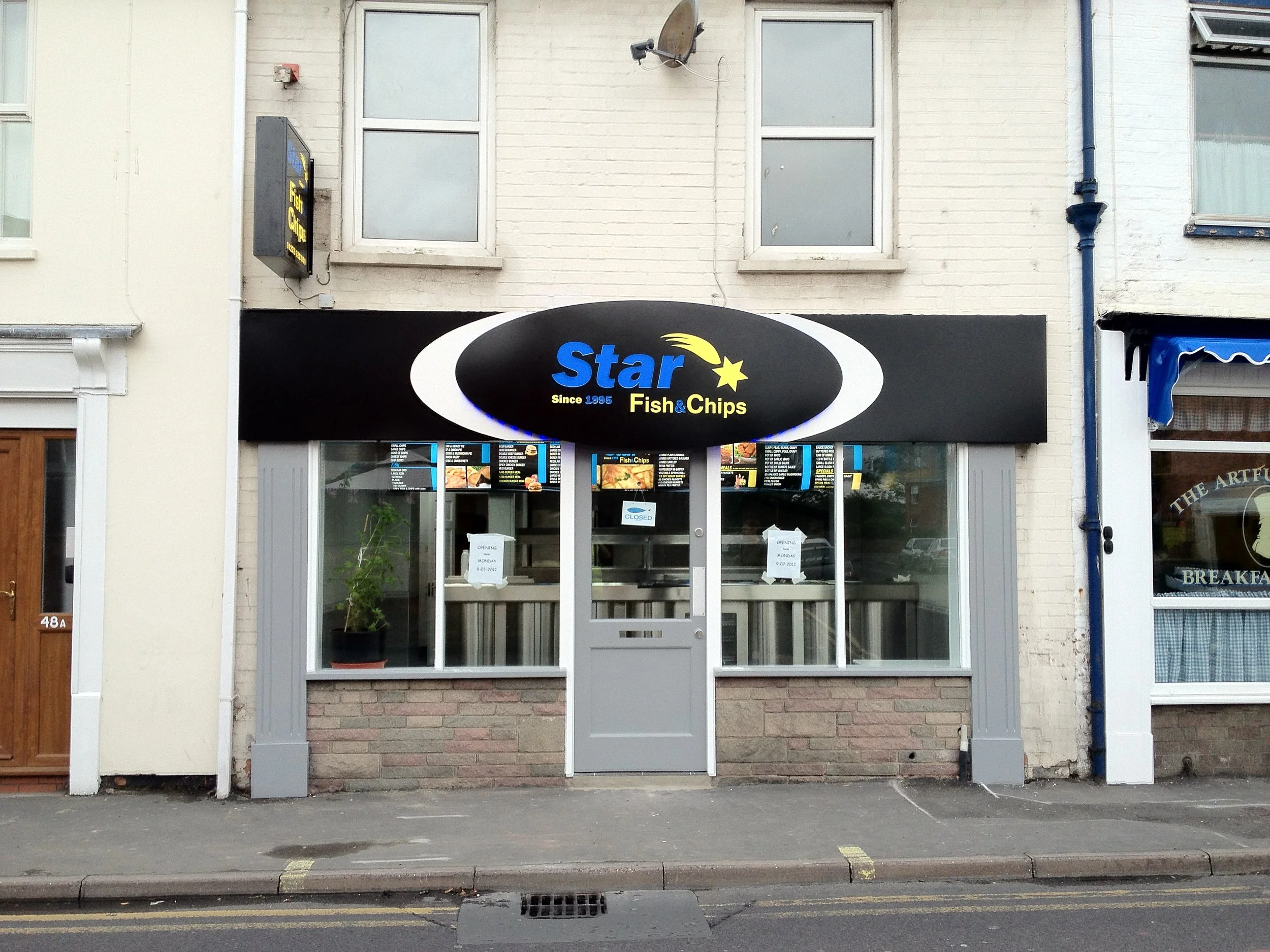 Storefront with black sign reading 'Star Fish & Chips' and digital menu boards visible through glass door, situated in a beige brick building with two upper windows, satellite dish, and a blue drainpipe.