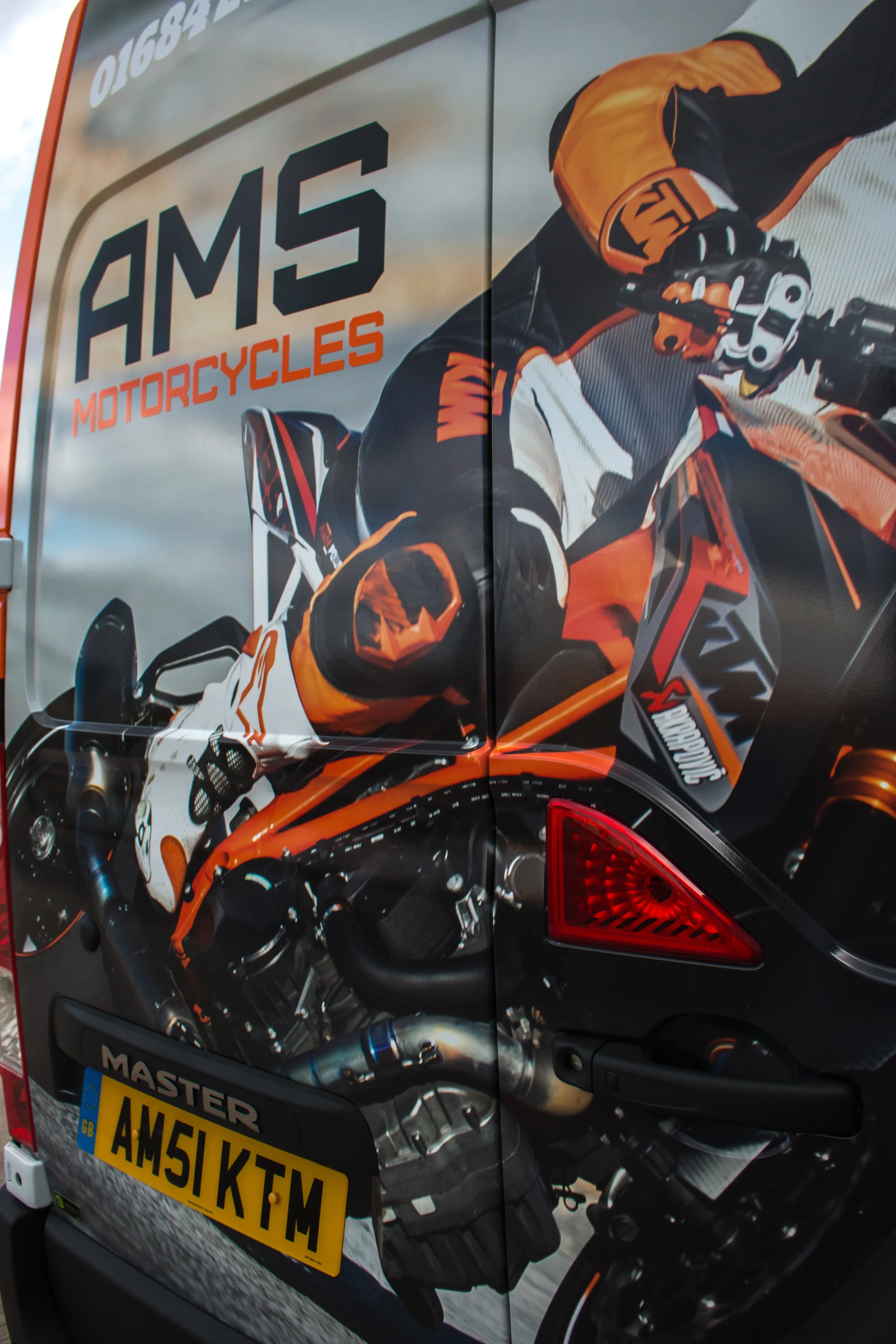 Close-up of a motorcycle with an advertisement on the vehicle's side showing a rider wearing orange and black gear on an orange motorcycle. The ad features the words 'AMS MOTORCYCLES' and a motorcycle image. The vehicle has a UK license plate reading