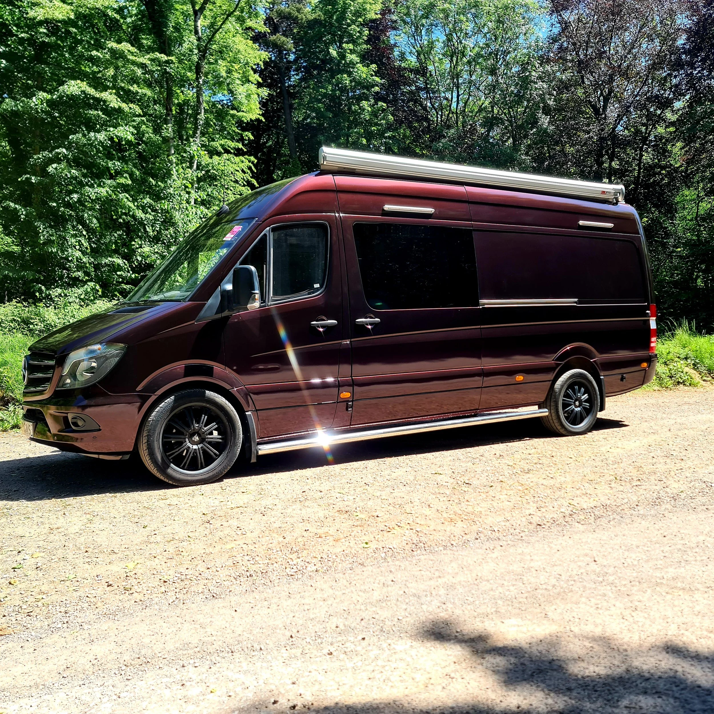 A maroon van parked on a dirt area with lush green trees and a clear blue sky in the background.