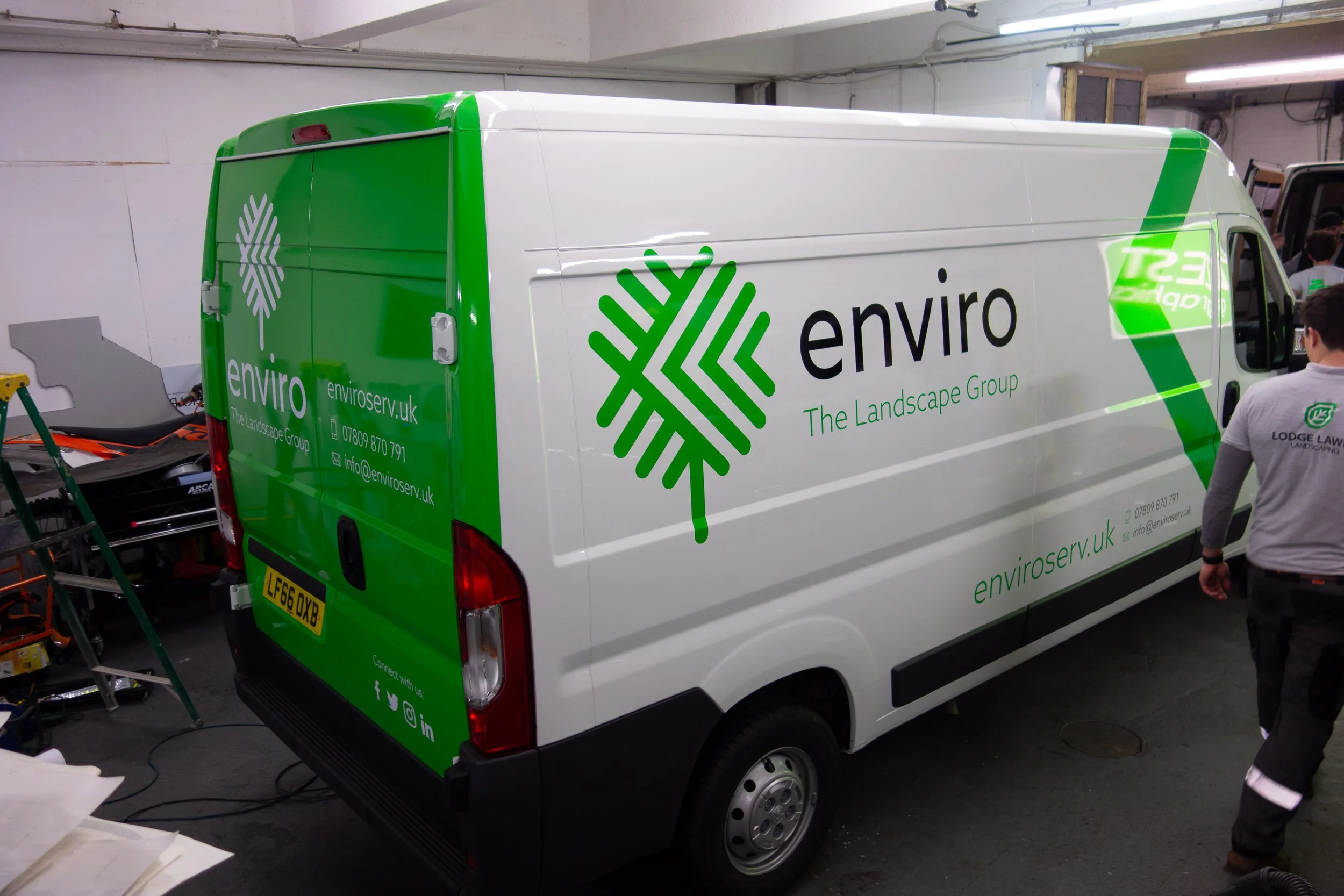 A white cargo van with green branding for Enviro, a landscape group, parked indoors. The logo features a green stylized tree and the website enviroserv.uk. There are people and equipment around, indicating it might be in a workshop or garage.