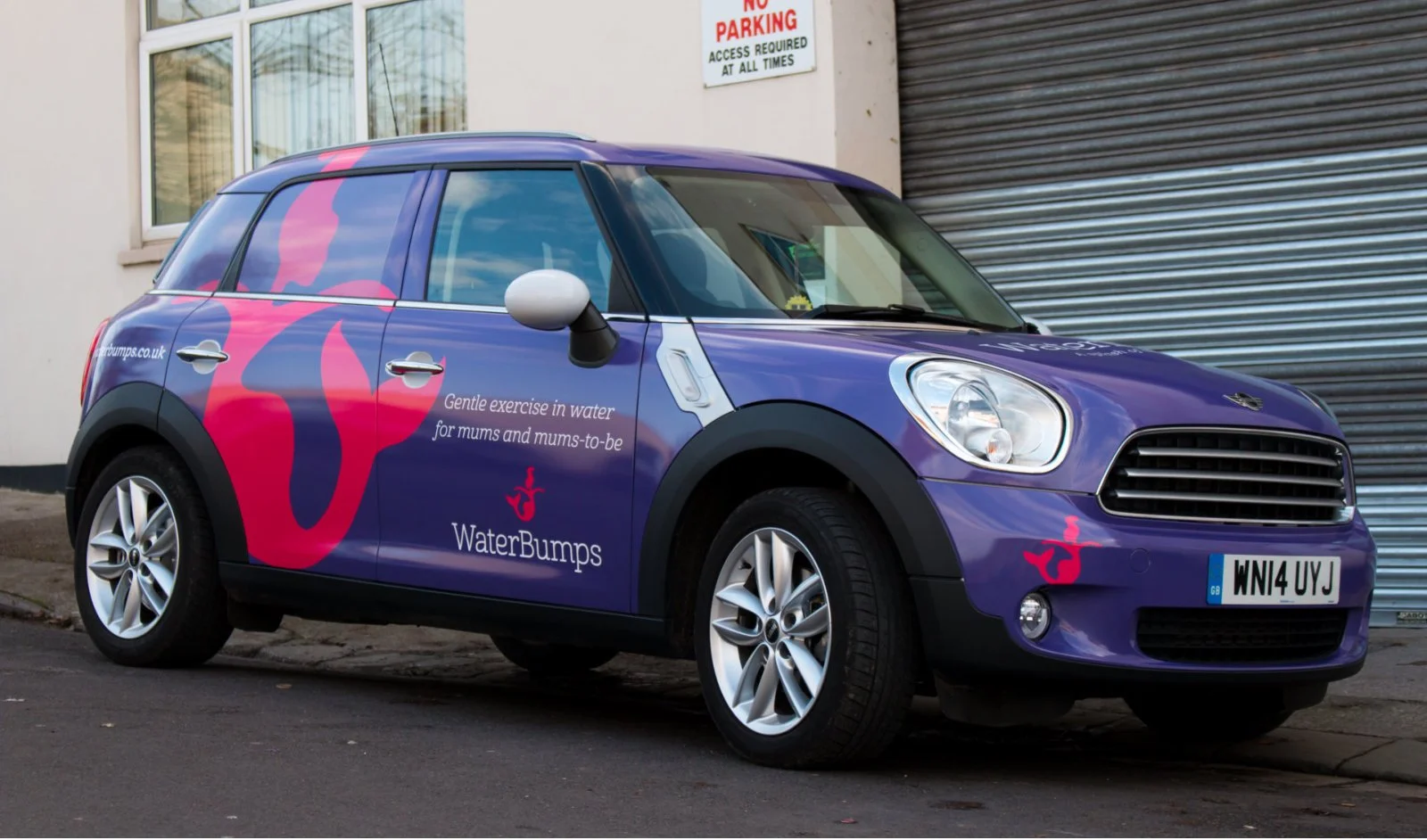 Purple compact car with pink water splash graphics and WaterBumps logo, parked on street next to a building with a "No Parking" sign, advertising gentle water exercise for mothers and expectant mothers.