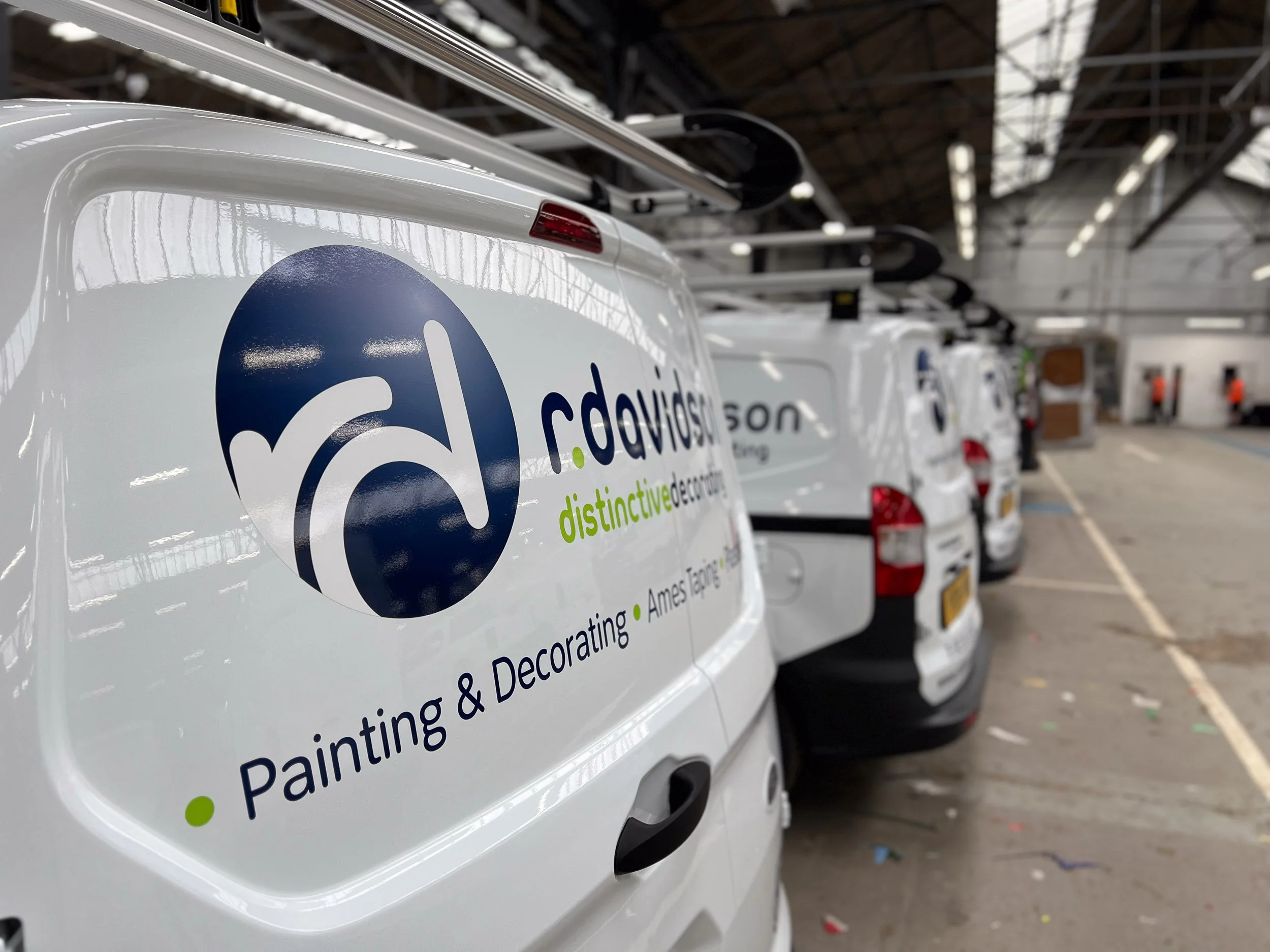 Close-up of the back of white vans with a logo and text related to painting and decorating in an industrial warehouse setting.