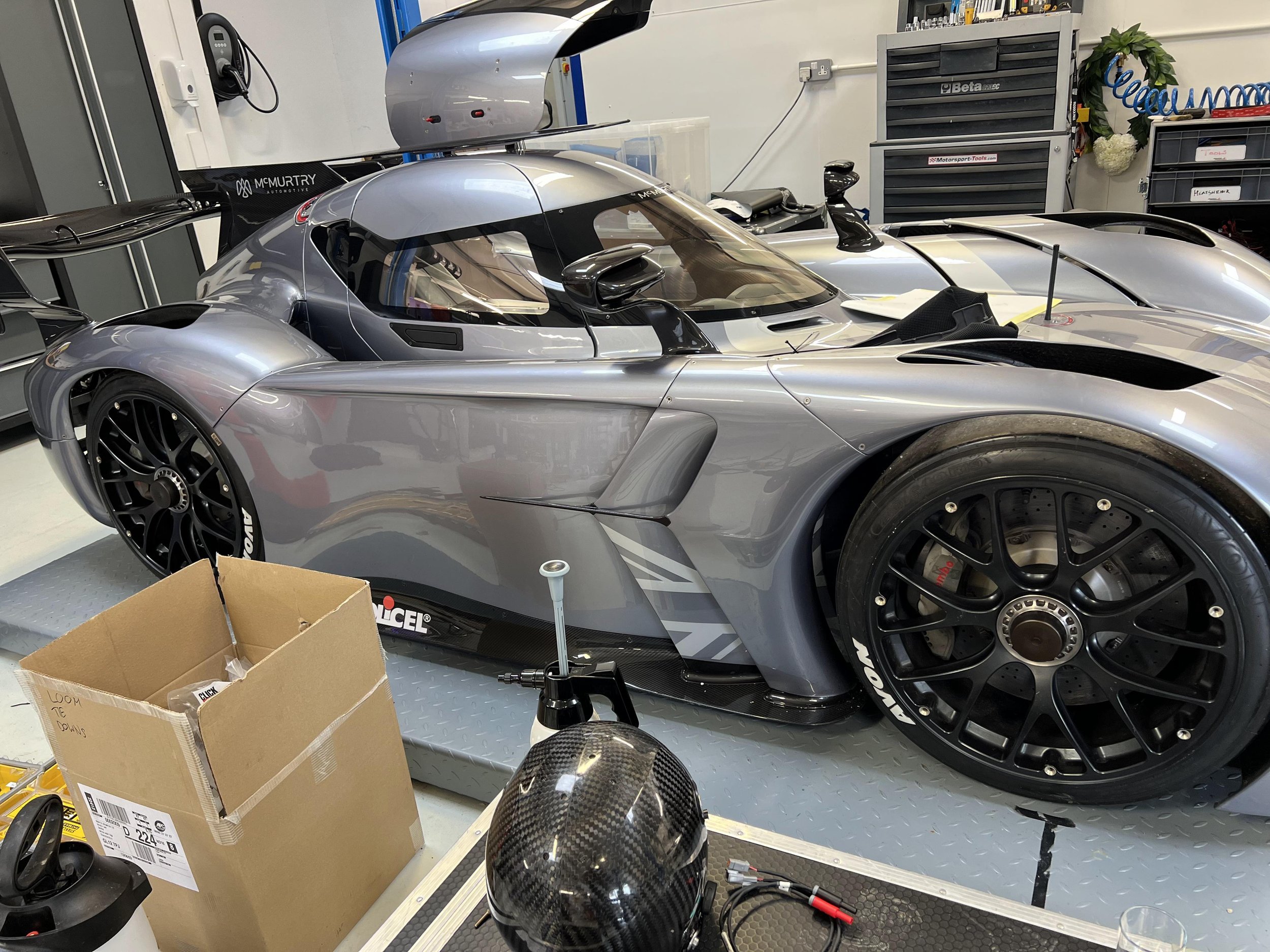 A silver and black race car in a workshop surrounded by various tools, equipment, and parts, including a cardboard box, a wheel, and fuel tank components.