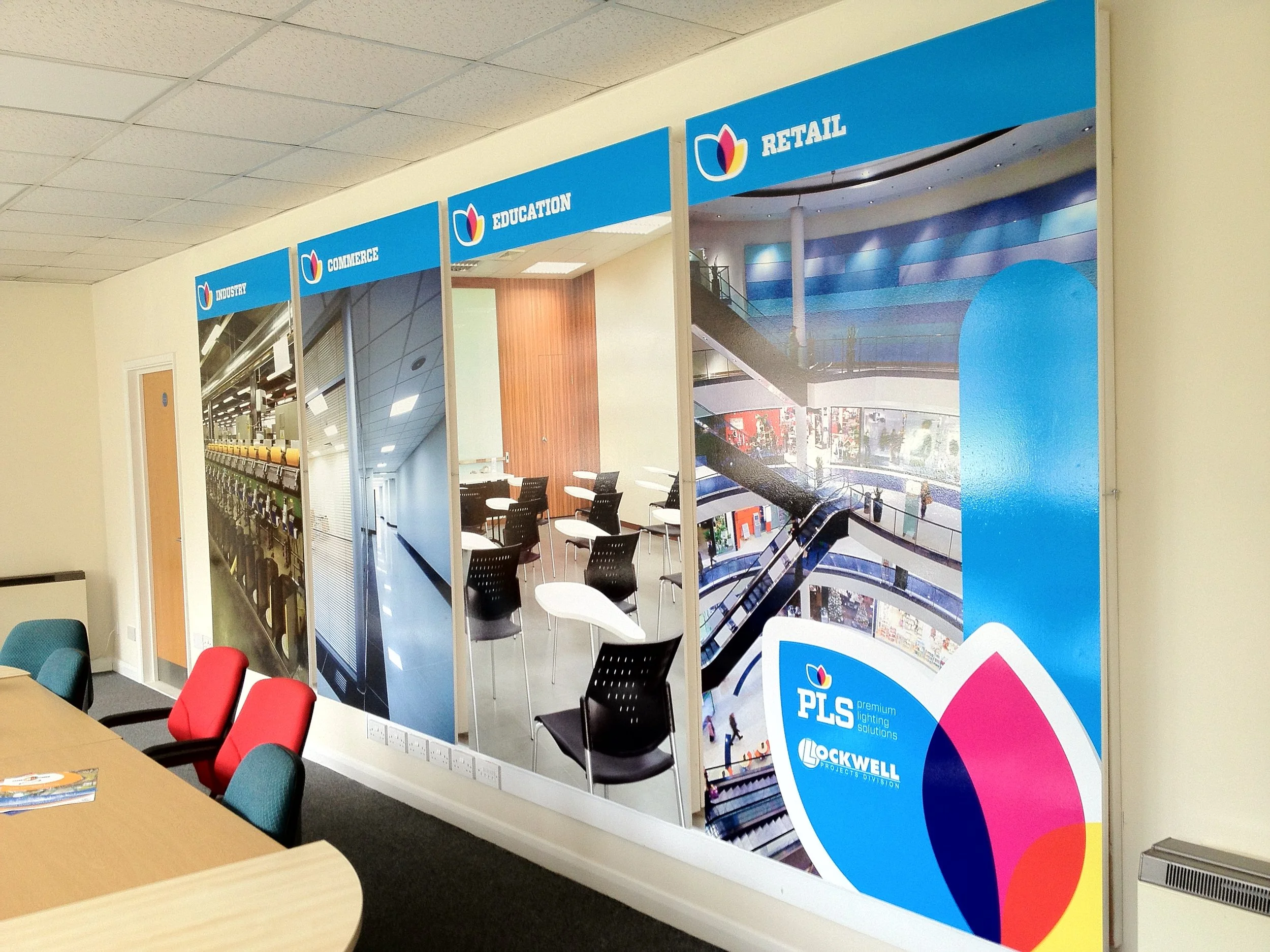 An office space with a wall display featuring images of shopping mall scenes and parking lot, labeled with categories 'Industry,' 'Commerce,' 'Education,' and 'Retail,' with seating and a table in the foreground.