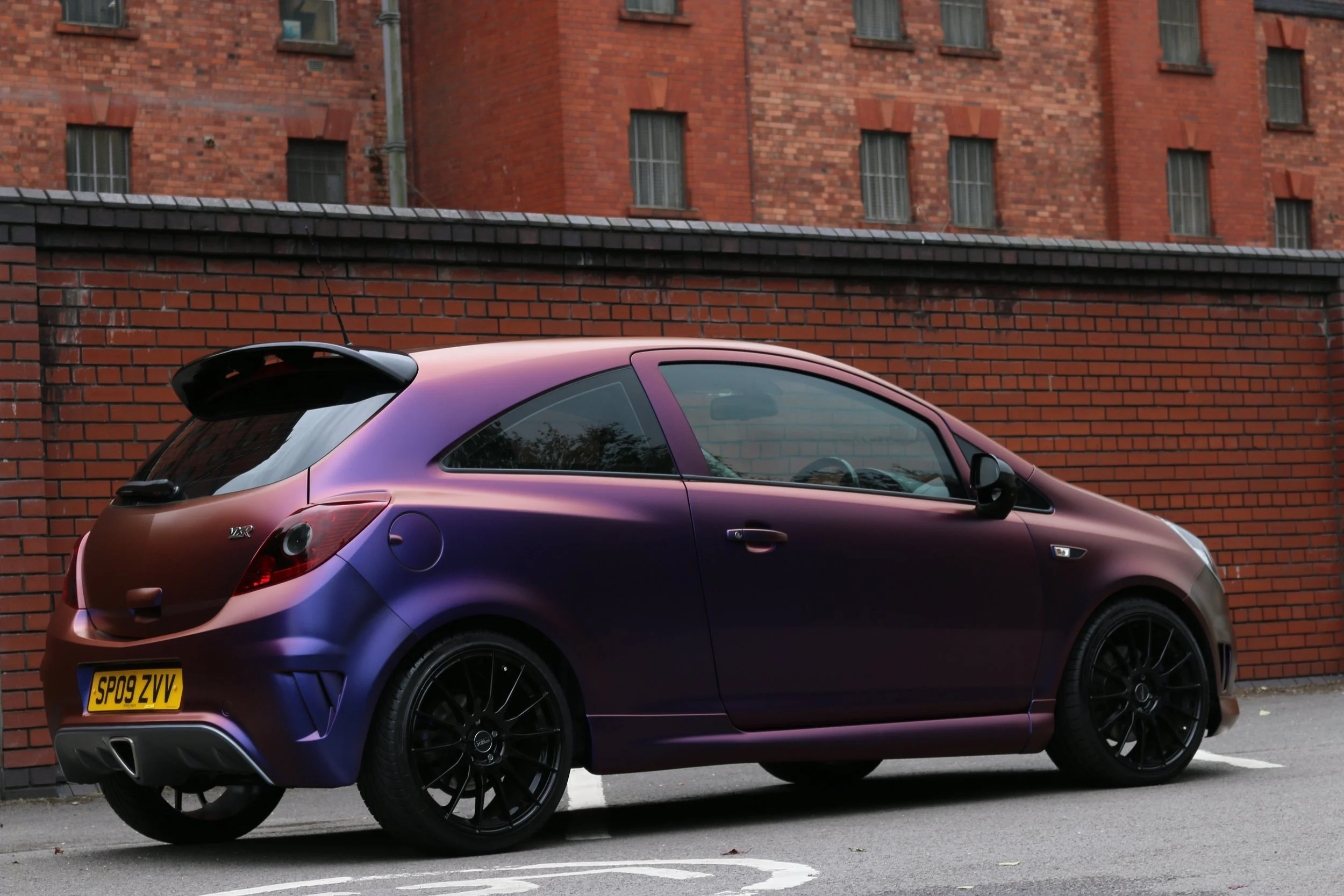 A purple hatchback car parked on a parking lot with a brick wall and building in the background.