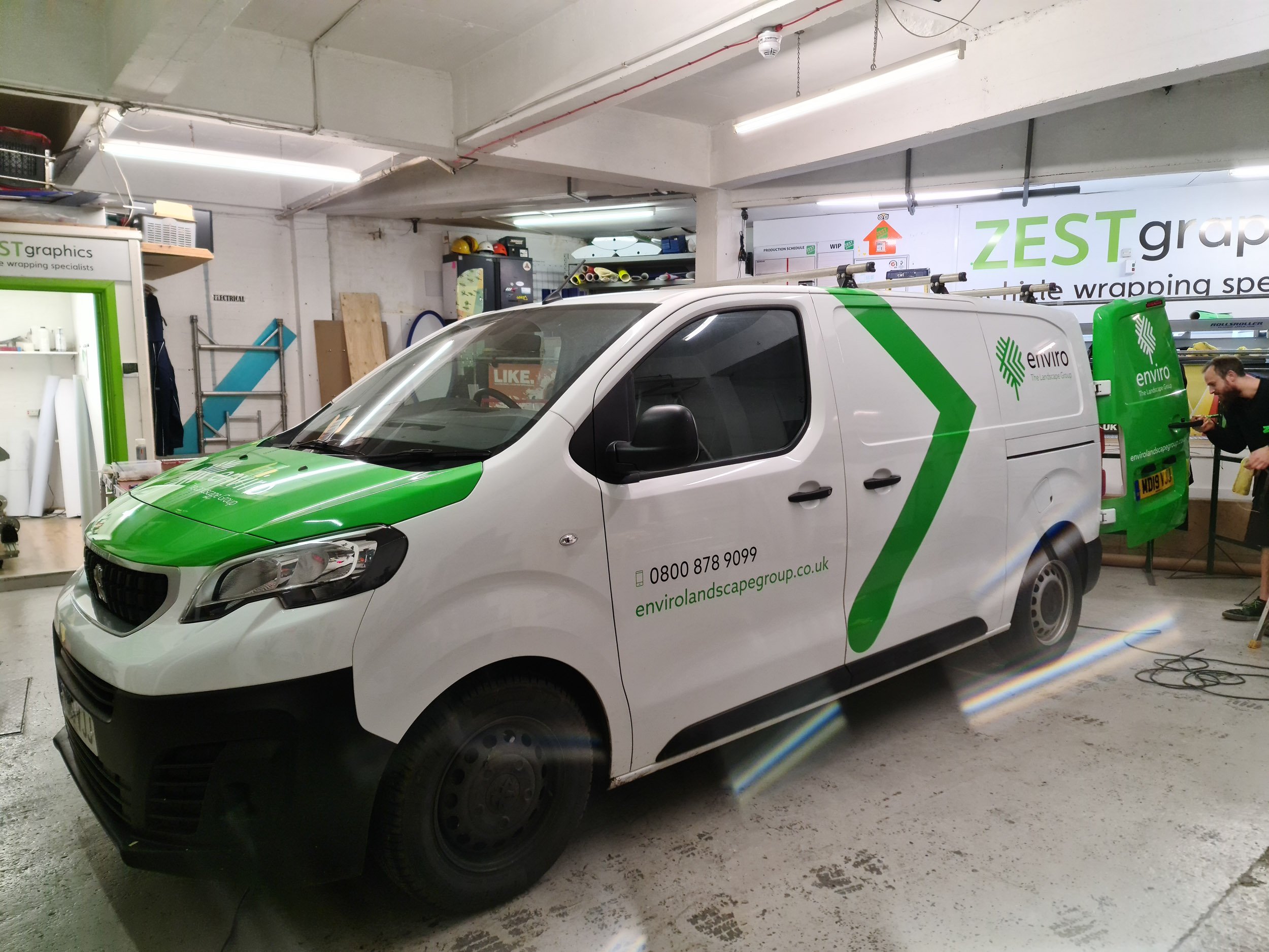 A white work van with green branding for Enviro is parked inside a warehouse or garage. The branding includes a leaf logo and the website envirolandscapegroup.co.uk. The van's hood and front are visible, and it has a black front bumper and black side
