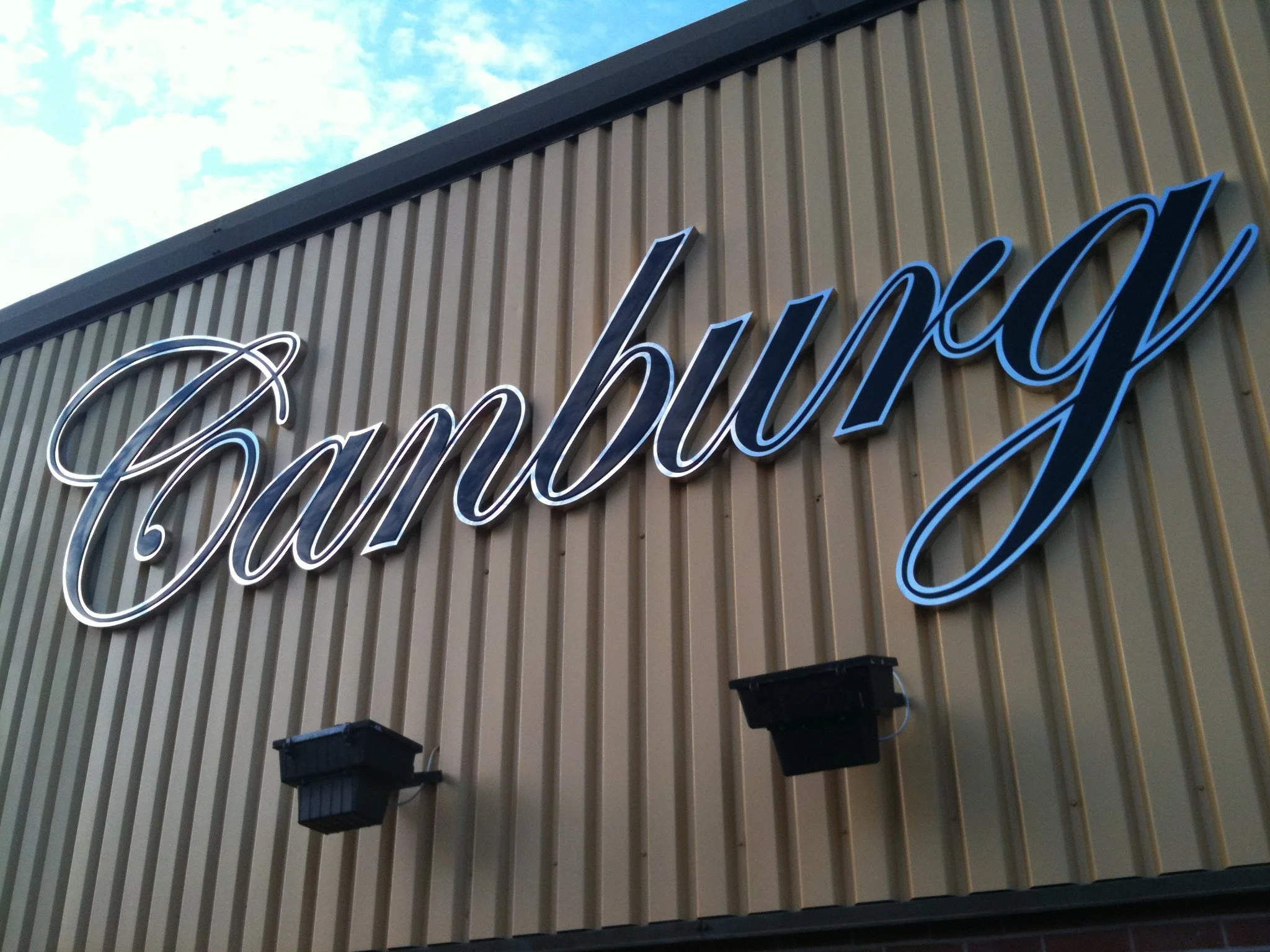Close-up of a building's exterior wall with vertical beige metal siding, featuring a large illuminated cursive sign that says 'Family' and two black wall-mounted planters below the sign.