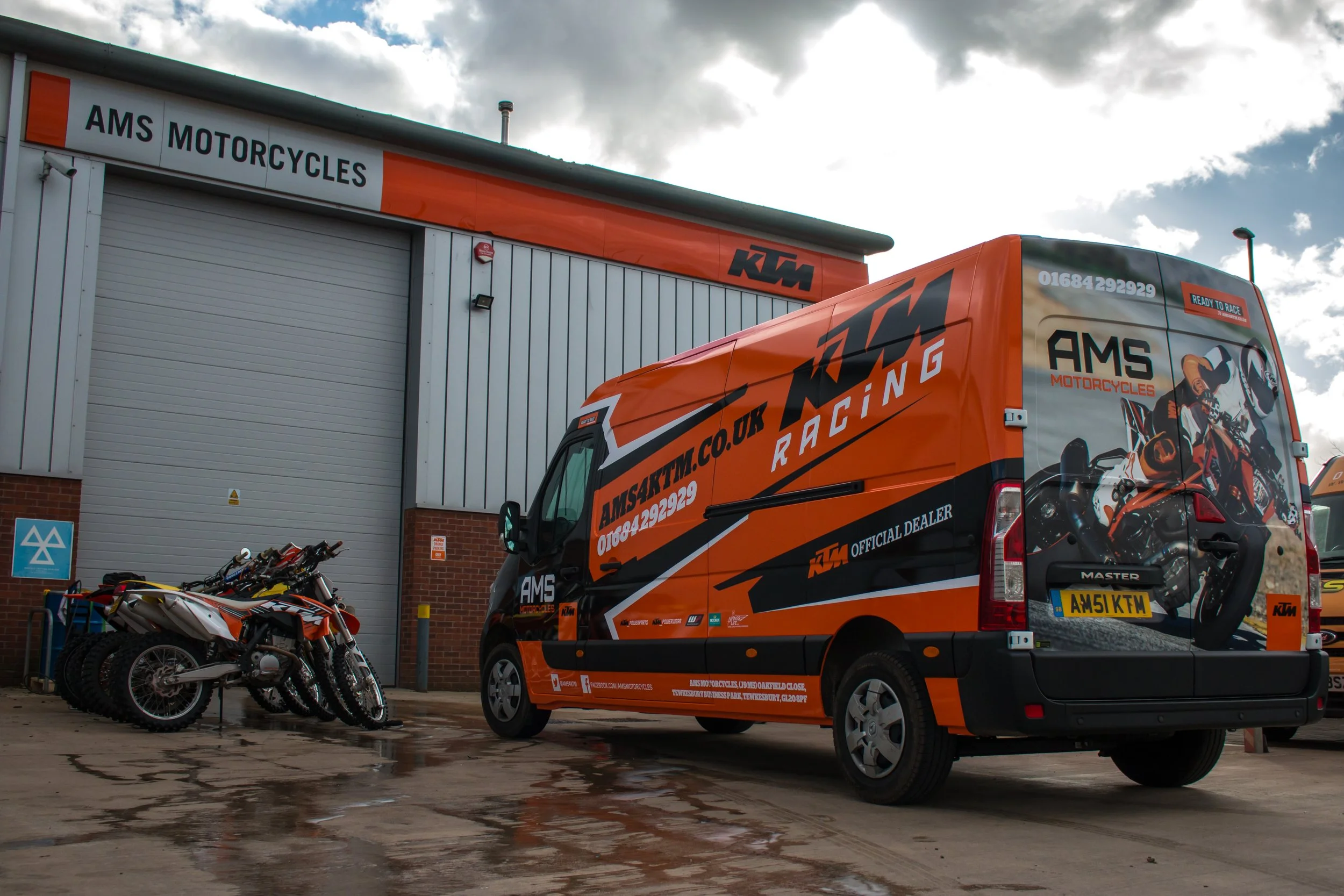 A KTM racing team support vehicle parked outside AMS Motorcycles shop, with dirt bikes lined up nearby on a wet pavement under cloudy skies.