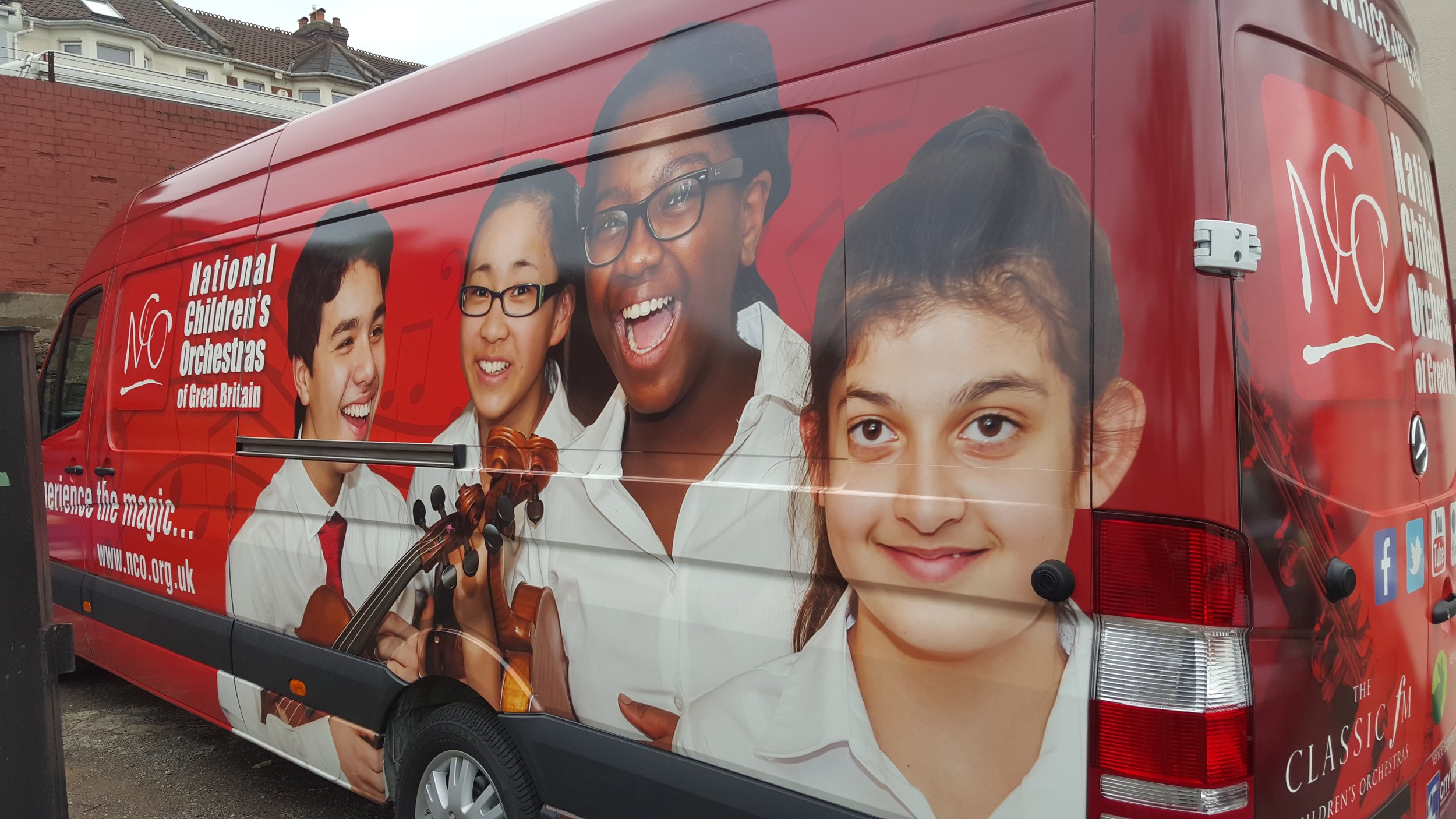 A red vehicle with a large wrap-around advertisement for the National Children's Orchestras of Great Britain, featuring images of children and a woman playing the violin.