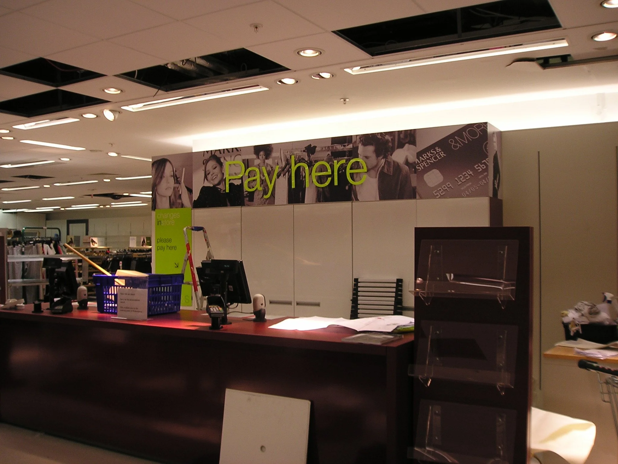 Check-out counter at a retail store with a sign that says 'Pay here' in bright yellow-green letters, with a background of promotional posters and store items.
