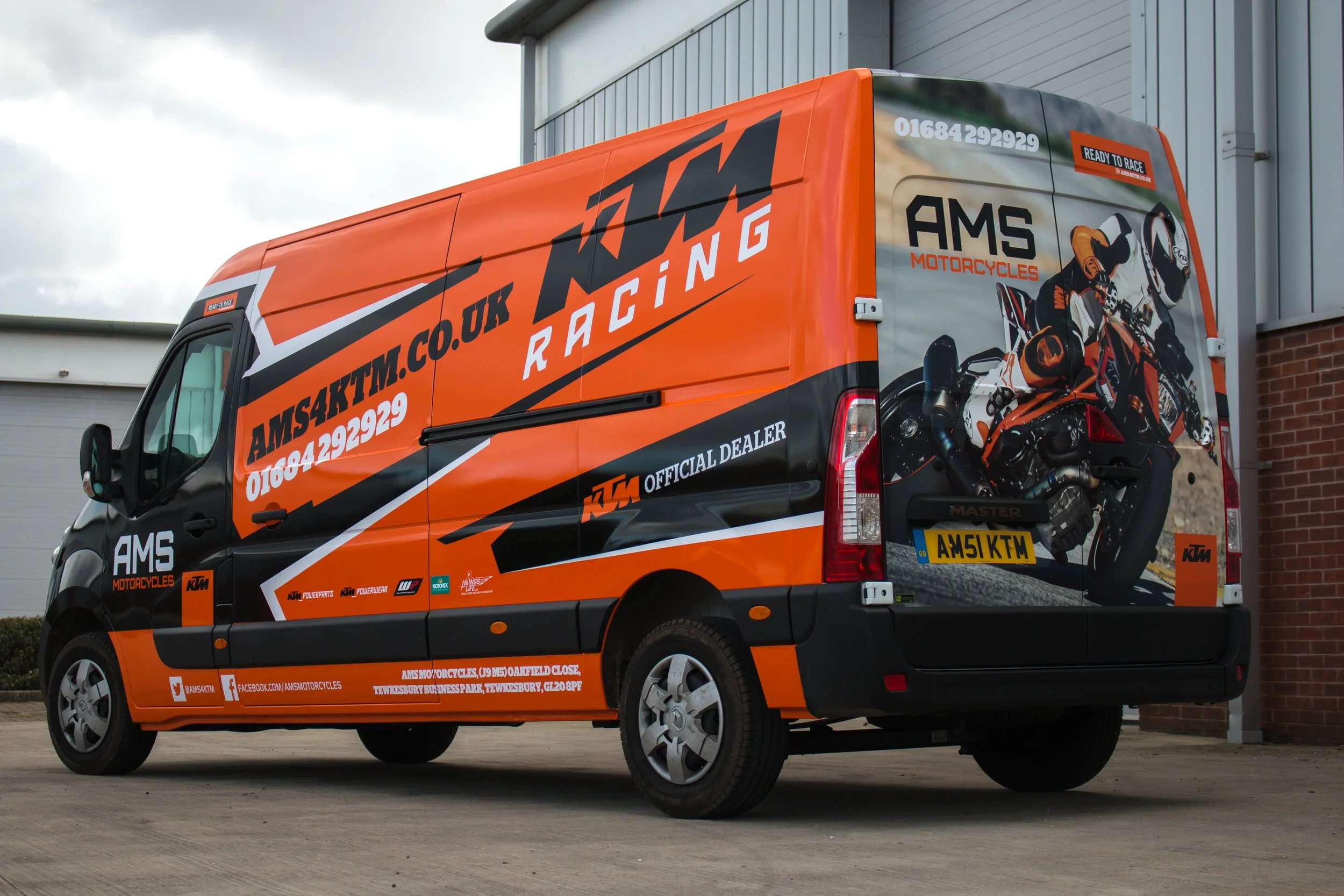 Orange KTM Motorcycles racing team support vehicle parked outside a building, displaying racing graphics and the team website on its side.