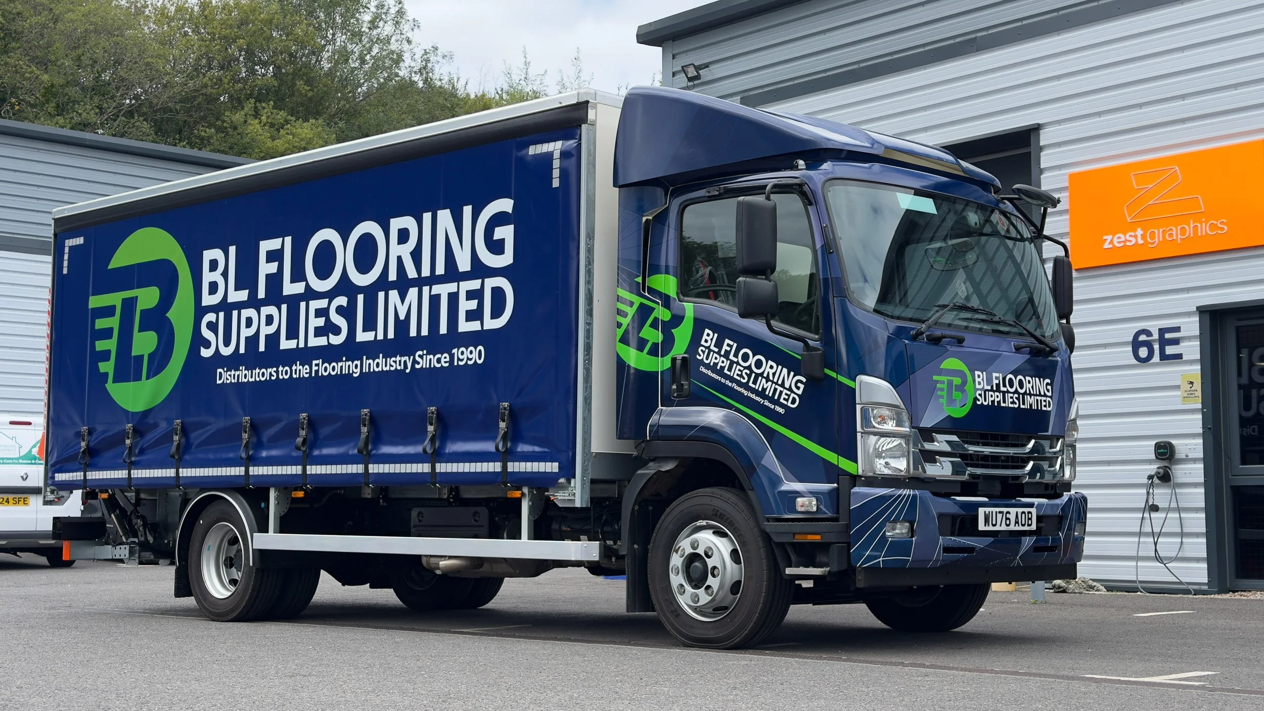 Blue delivery truck with green and white lettering for BL Flooring Supplies Limited parked outside a commercial building.