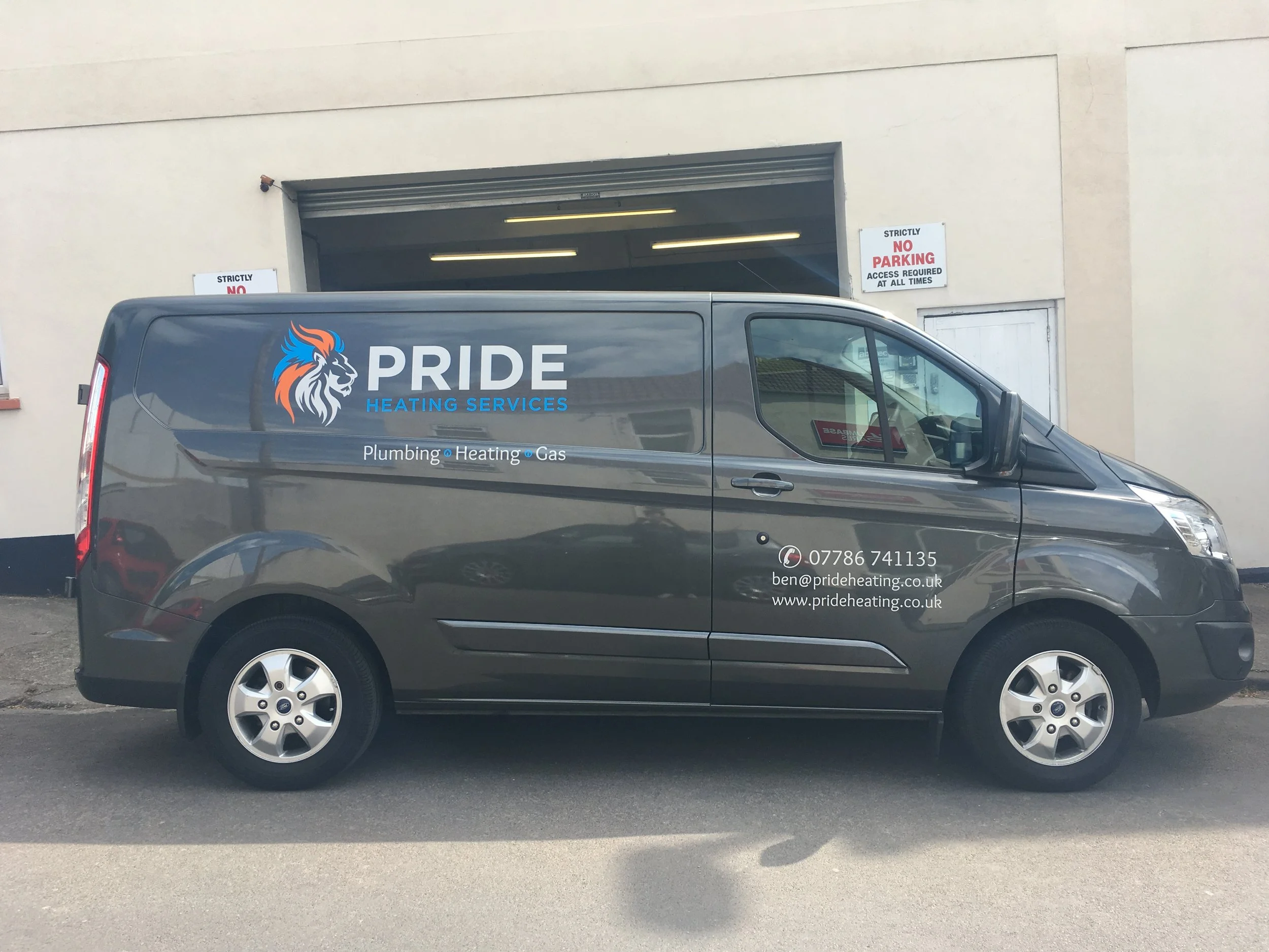 A company van for Pride Heating Services parked outside a building with a large open garage door. The van is dark gray and features the company logo, which includes a stylized lion with a blue mane, and text advertising plumbing, heating, and gas ser
