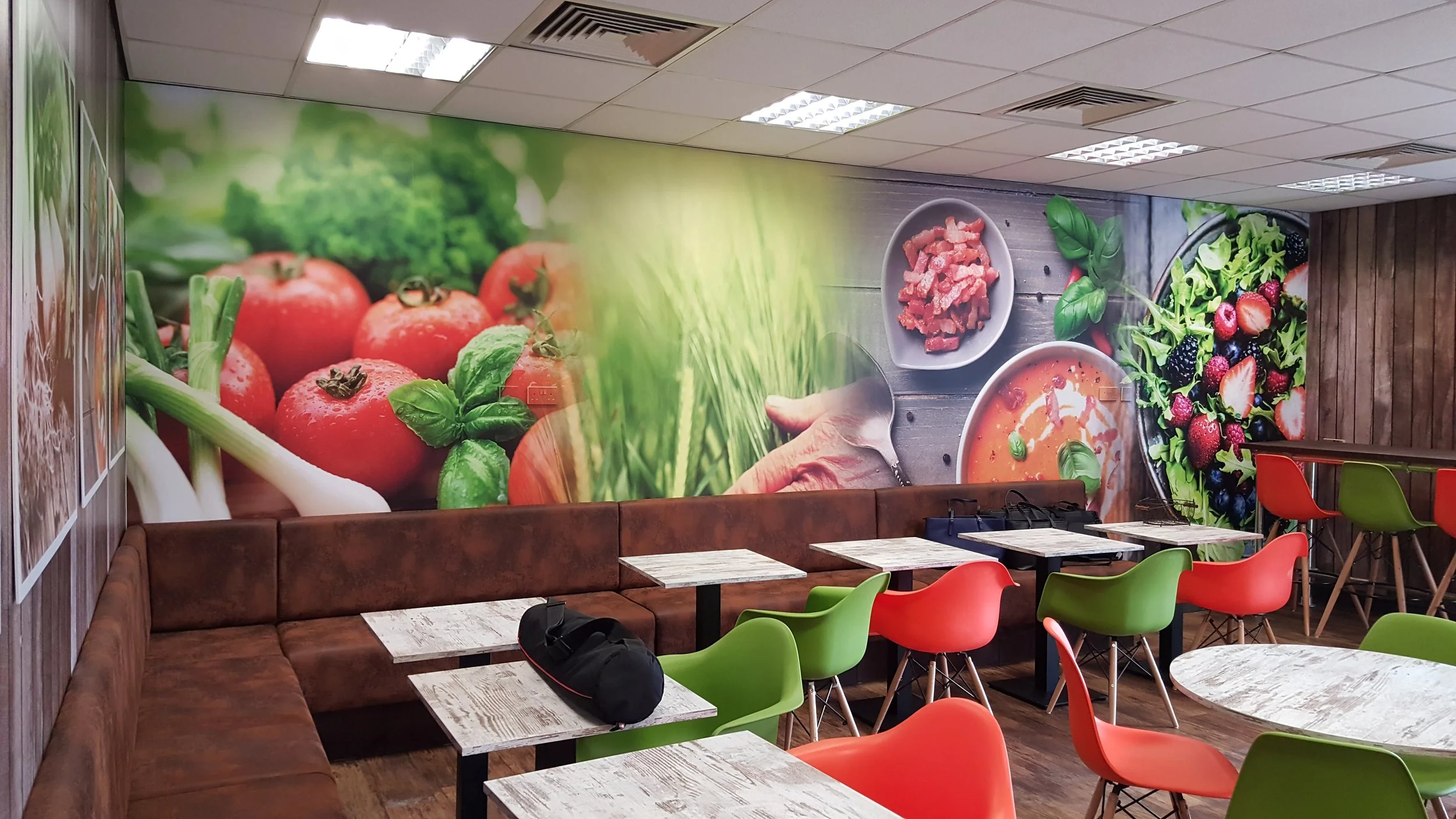 cafe interior wall digitally printed wall wrap mural featuring a close-up of fresh tomatoes, green onions, basil leaves, and bowls of sliced meat, soup, and a mixed berry salad, with tables and colorful chairs in the foreground.