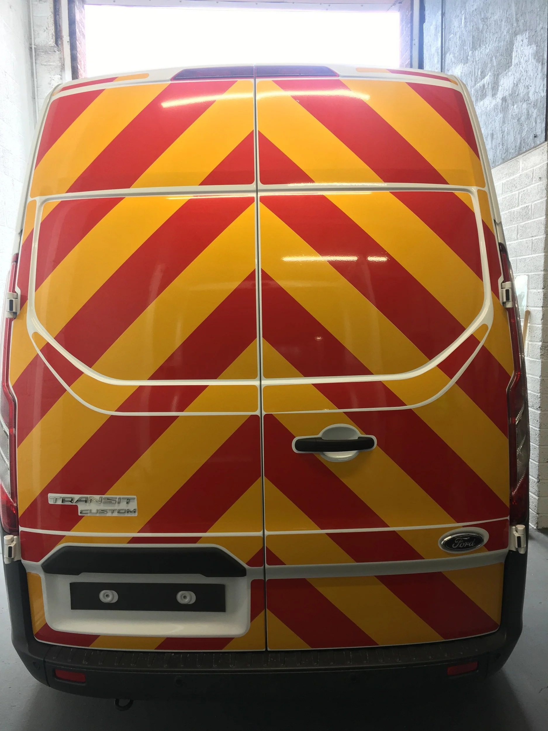 Rear view of a white Ford Transit Custom van with red and yellow diagonal stripe safety markings on the back doors, parked in a garage.