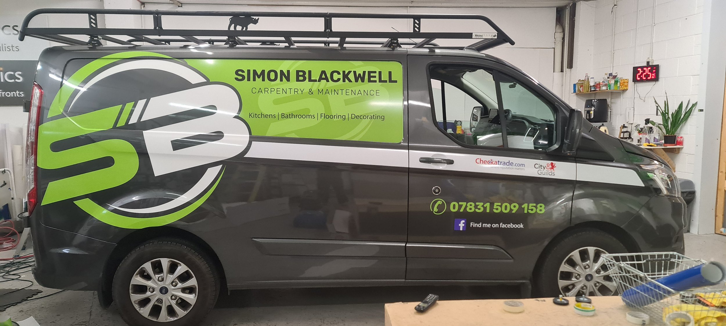 A commercial van with branding for Simon Blackwell Carpentry & Maintenance, parked indoors. The van is gray with a large green and white logo and text listing services and contact information, including phone number and Facebook. The background shows
