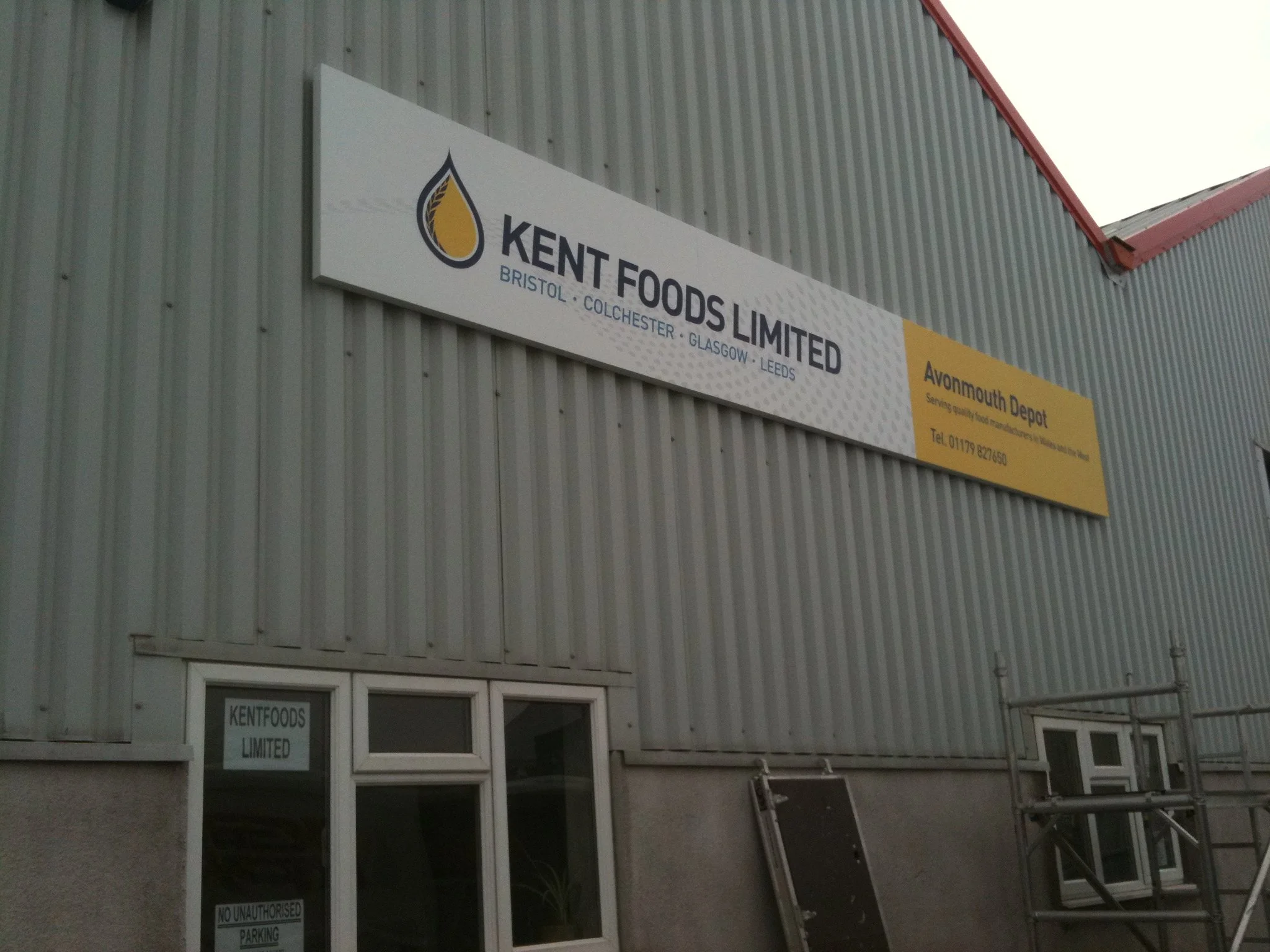 Exterior of a building with a sign reading 'KENT FOODS LIMITED' and locations listed as Bristol, Colchester, Glasgow, Leeds. A smaller sign below the main sign indicates the location as Avonmouth Depot. The building has striped metal siding and a red