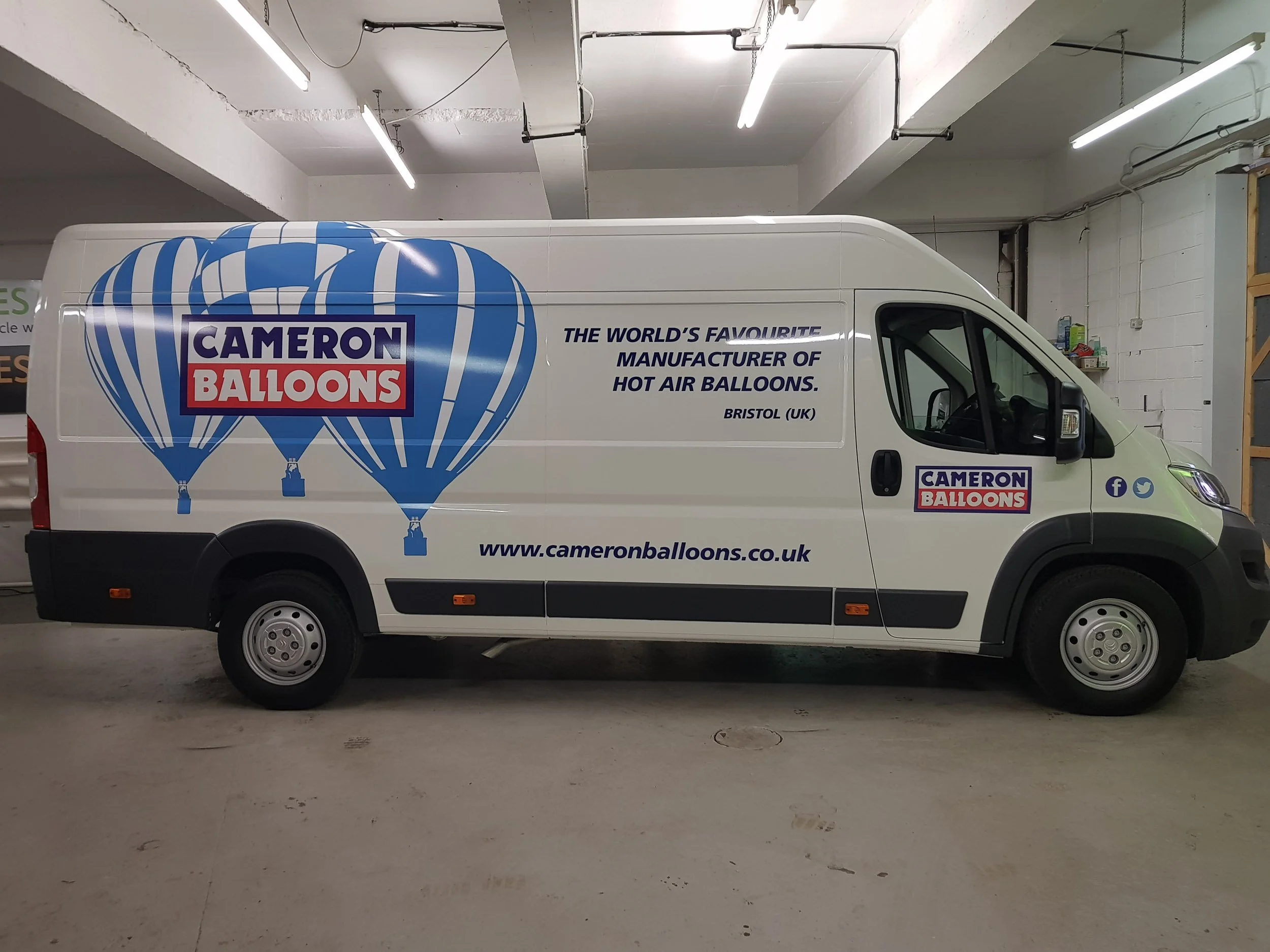 A white cargo van with the Cameron Balloons logo and slogan, parked inside a warehouse with fluorescent lighting and shelves.