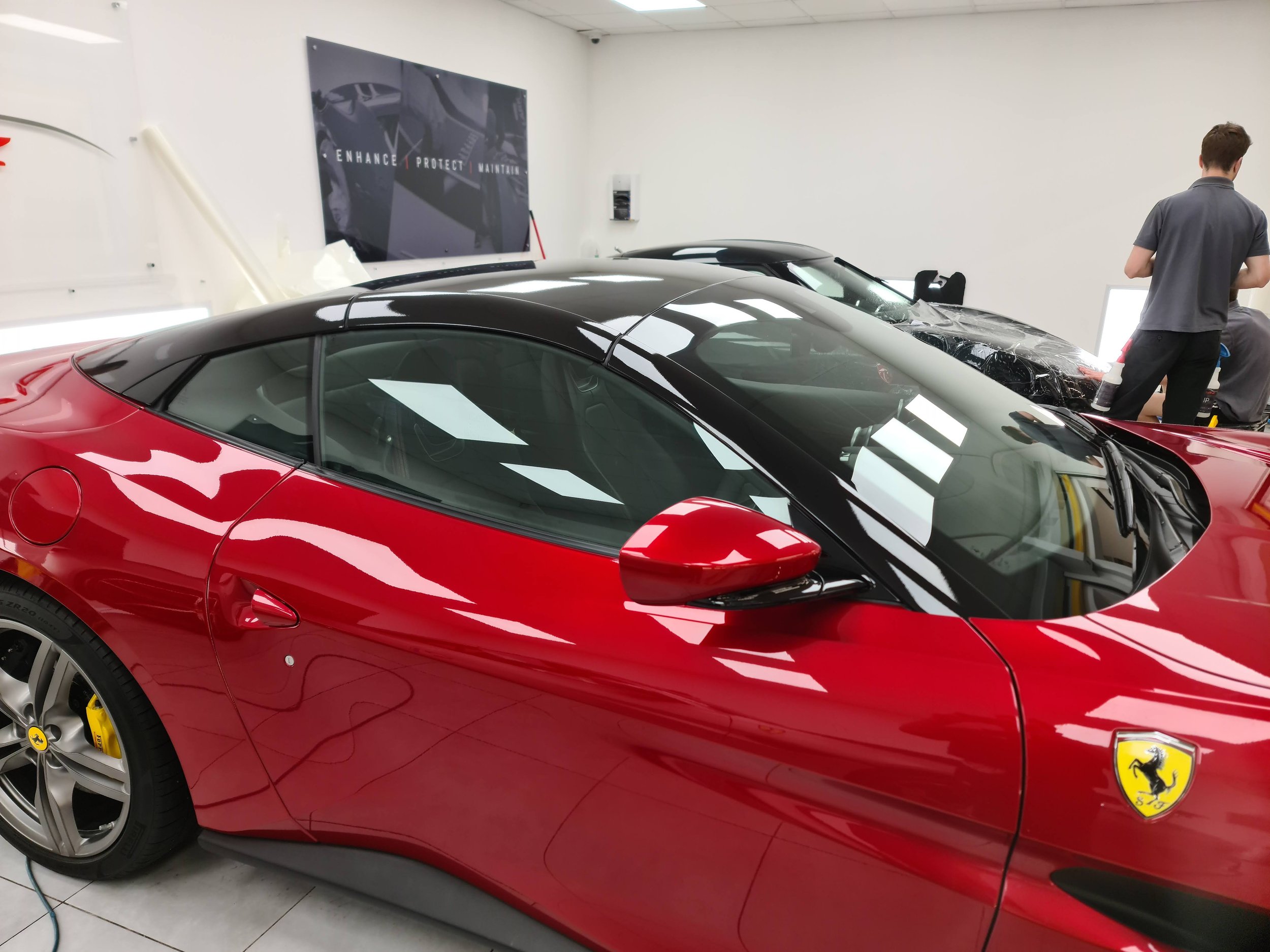 Inside a car dealership, a red sports car with black roof and yellow brake calipers is parked beside several other sports cars. Two people are working on a silver car in the background. A poster on the wall reads 'Enhance. Protect. Maintain.'