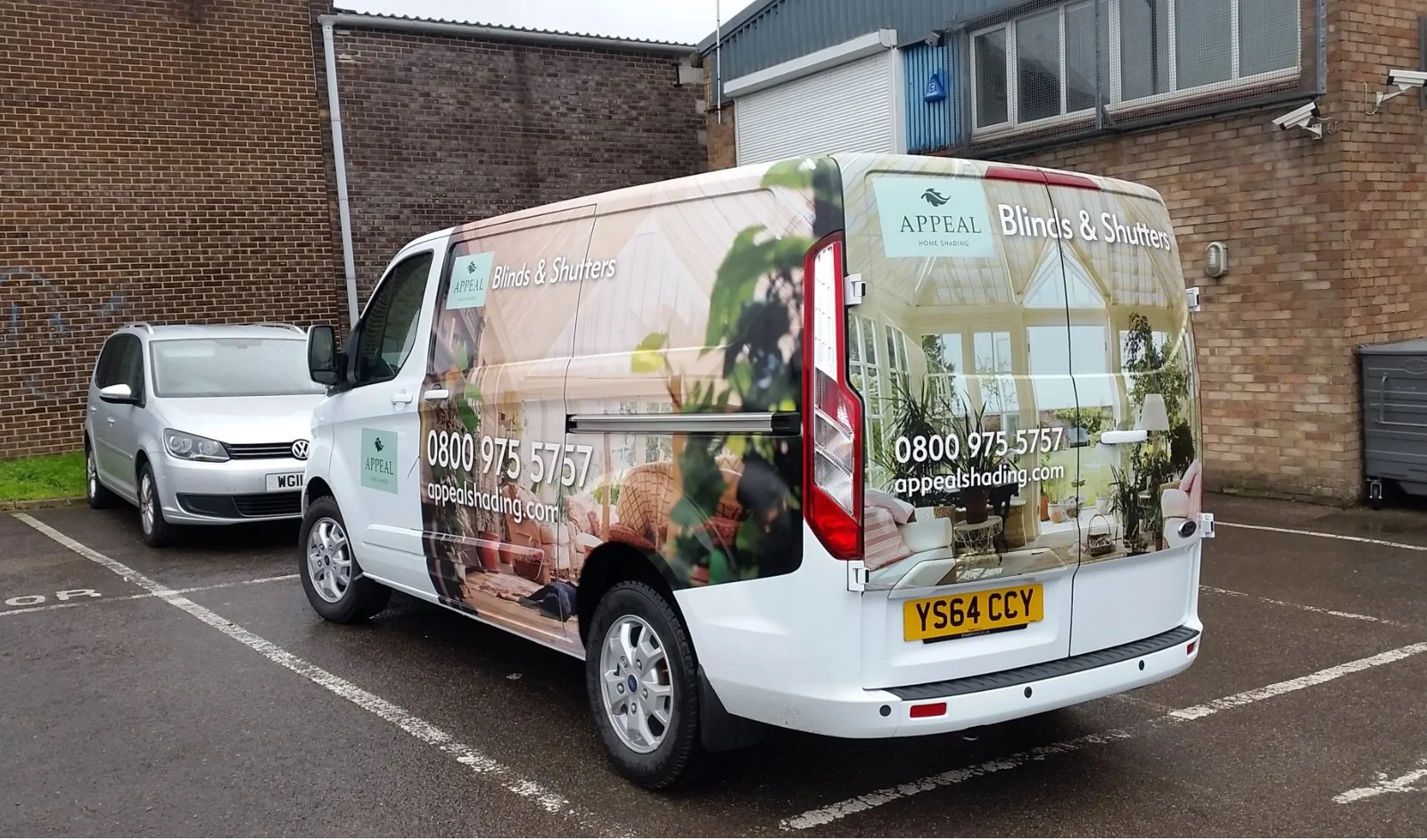 A white commercial van parked in a parking lot, advertising for a company called Appeal that specializes in blinds and shutters, with contact information and a website listed, with two other cars and a brick building in the background.