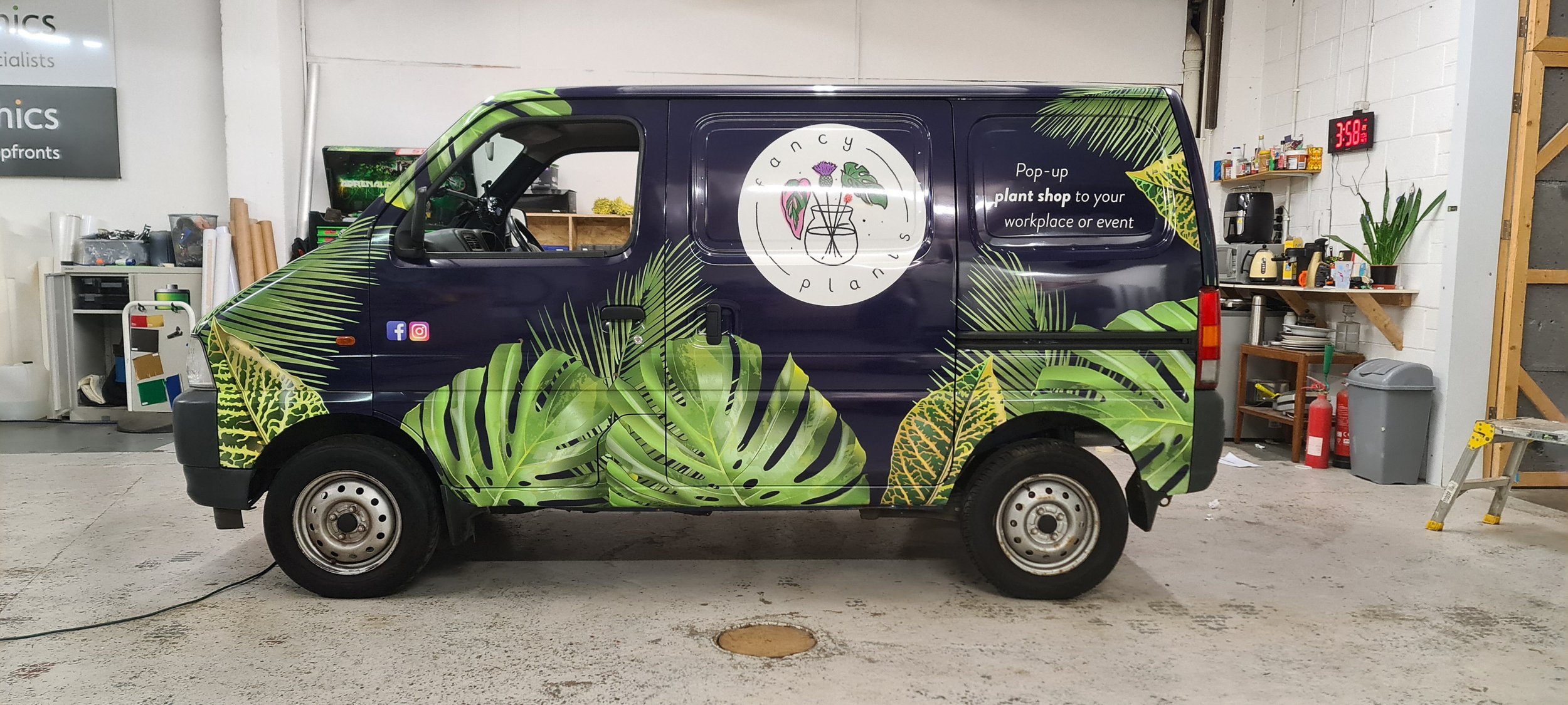 A van with a dark blue base color featuring a large green tropical leaf pattern on the sides. The van has a round logo on the side that reads 'FANCY PLANTS' with an illustration of a plant in a pot surrounded by various leaves and flowers. The text o
