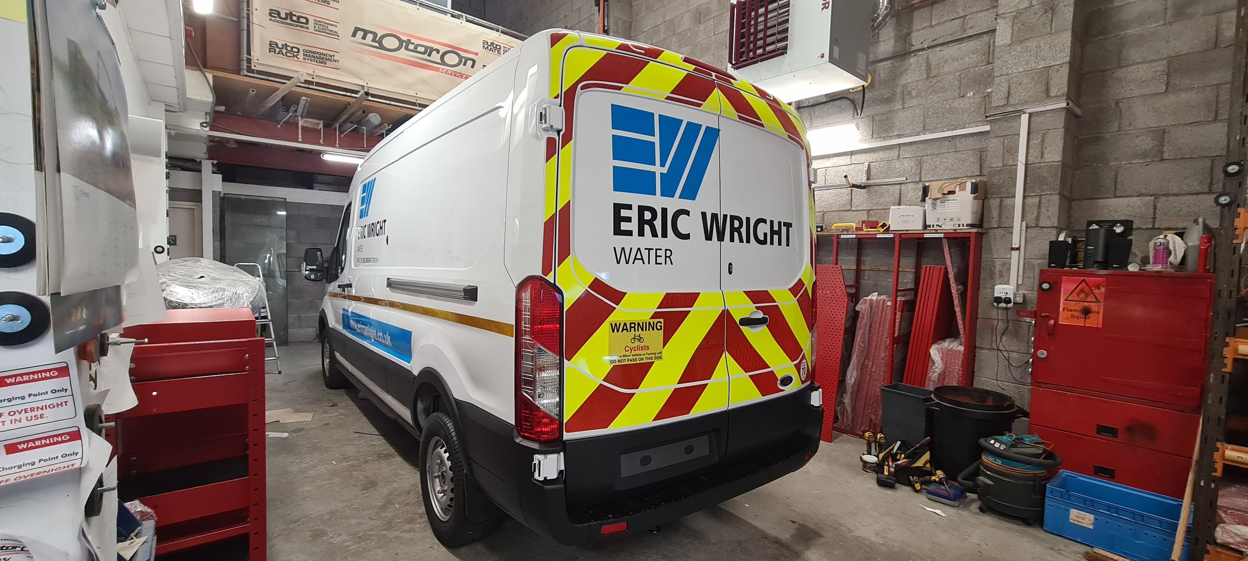 Water delivery van in a garage with various tools and equipment, including red tool chest, shelves, and warning signs.