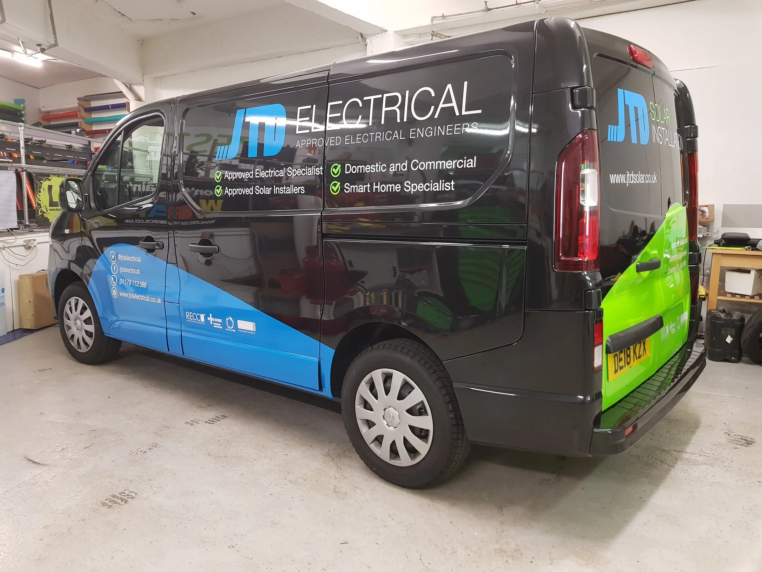 A black commercial van with blue and green graphics and text advertising JD Electrical and Solar Installation, parked indoors on a concrete floor.