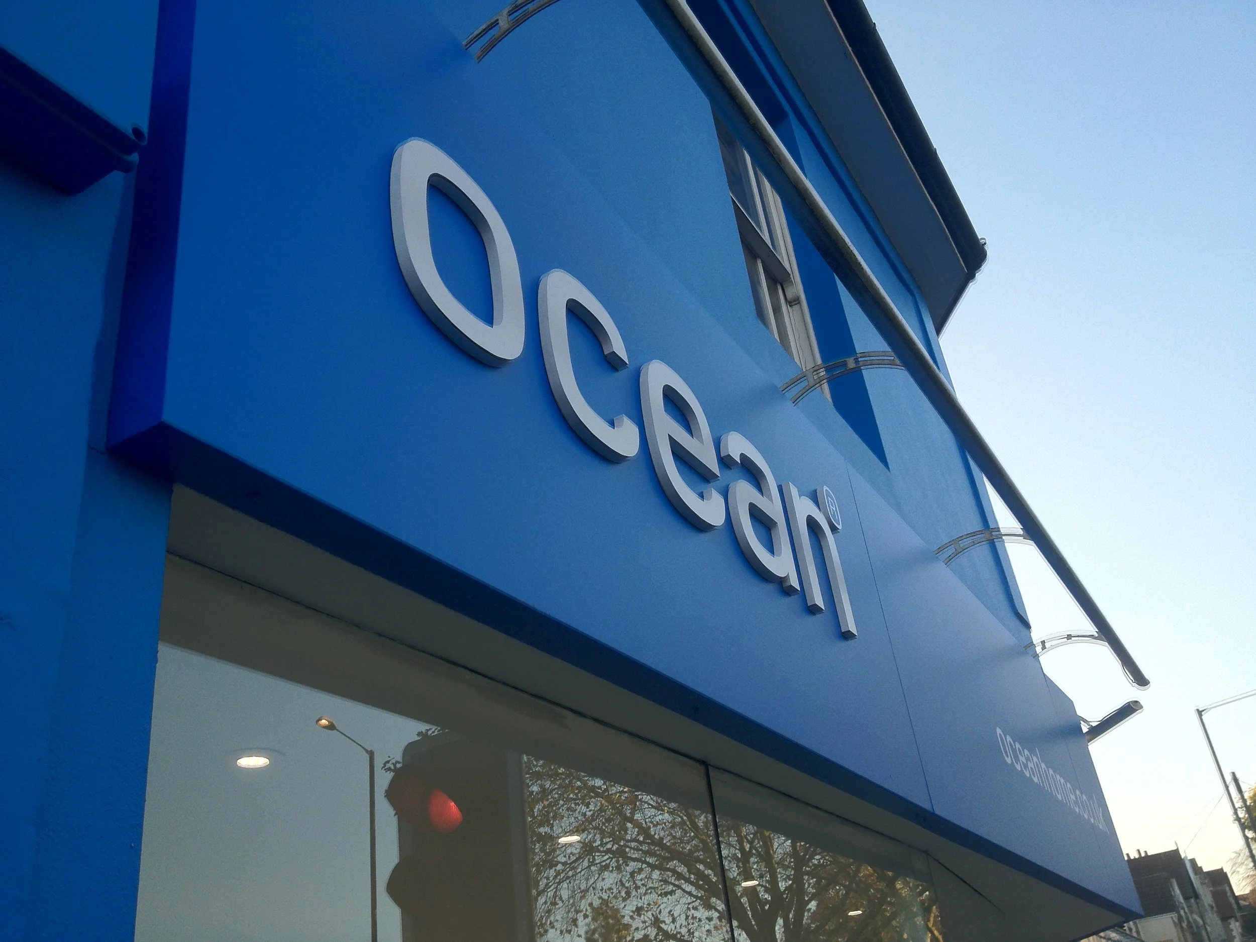 Close-up of a blue building exterior with the white lowercase text 'oc' visible, indicating a sign for a business or organization, reflecting on a nearby glass surface.