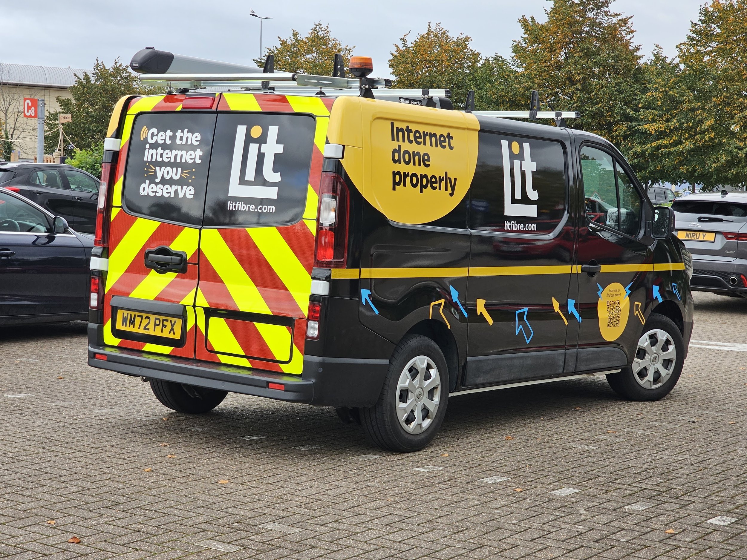 A black and yellow service van parked in a parking lot, fully wrapped in gloss black with slogans promoting internet service and a website address, with trees and other vehicles in the background.