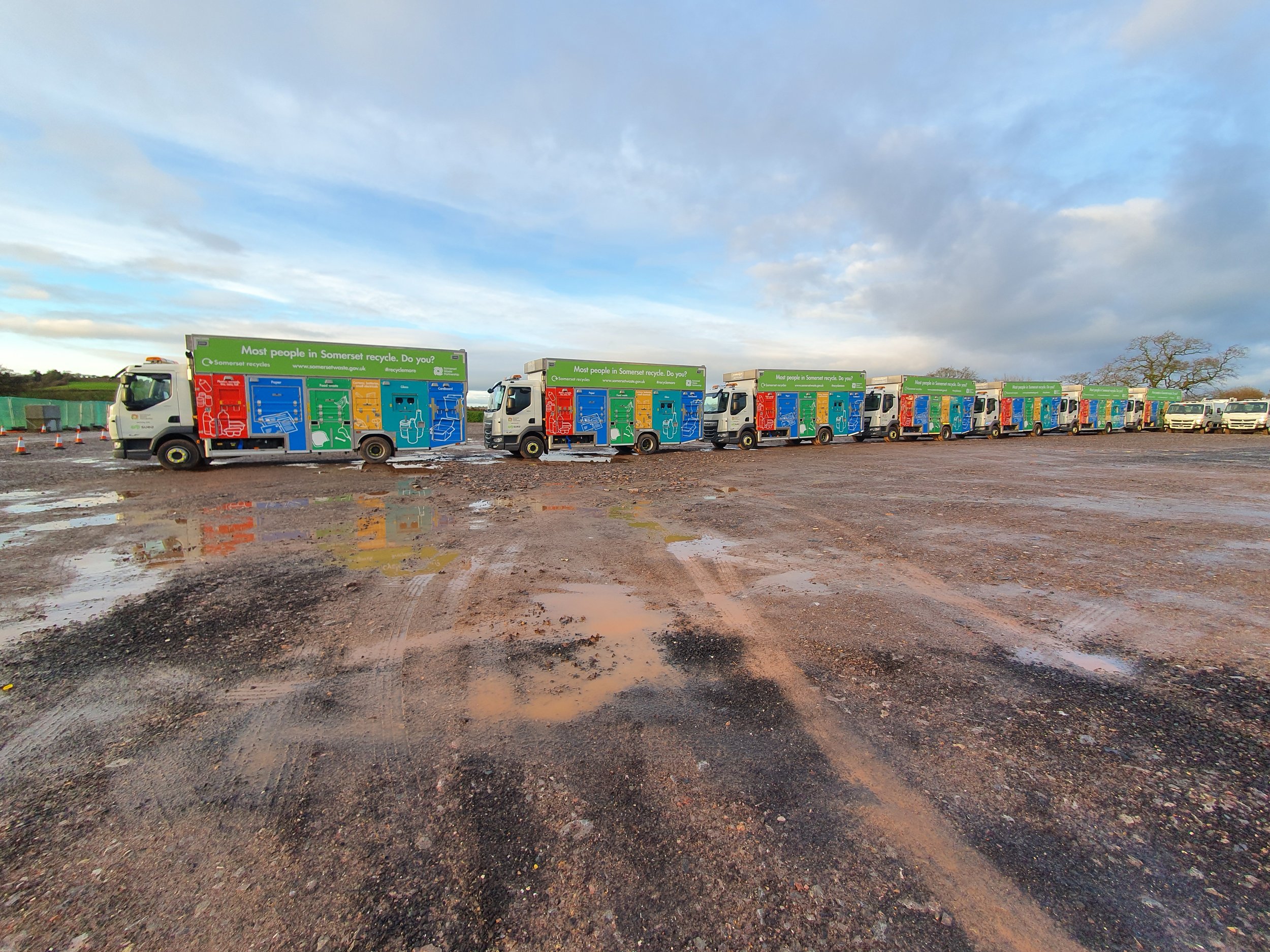 Line of recycling trucks with colorful graphics and a message about recycling, parked on a muddy surface with puddles, under a partly cloudy sky.