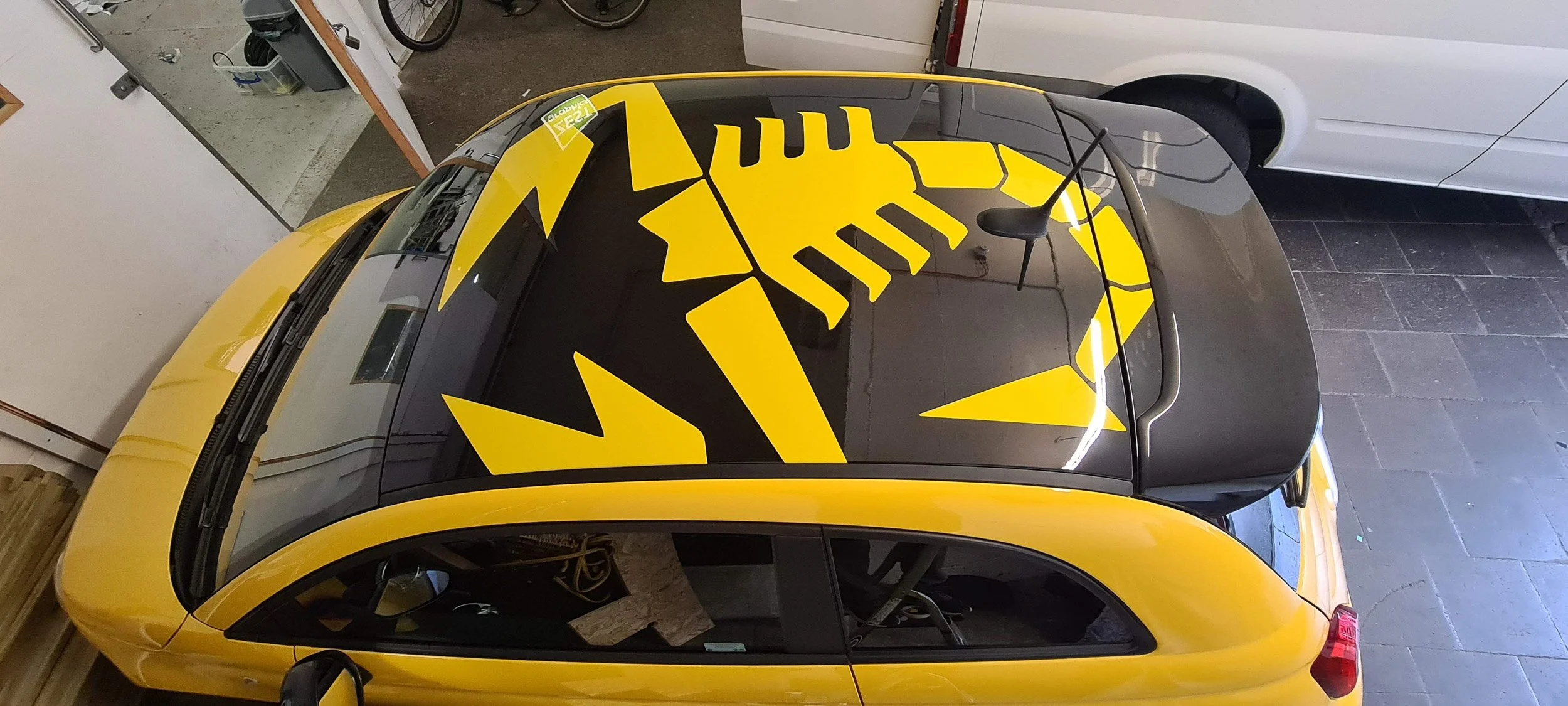Yellow and black sportscar with graphic design on the roof, parked inside a garage with bicycles and other items nearby.