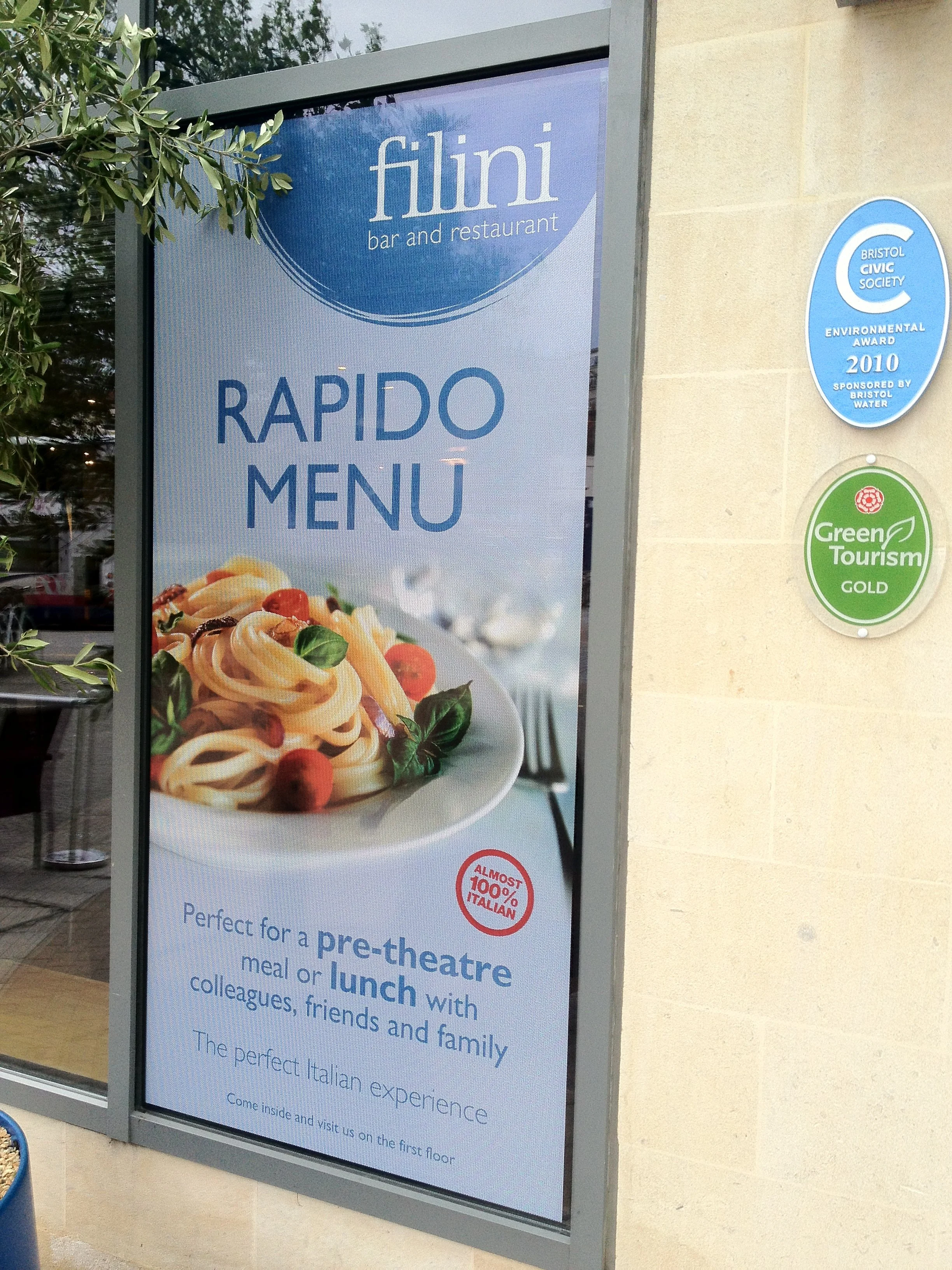 Digital sign outside a restaurant with the name 'Filini bar and restaurant', displaying a 'Rapido Menu' with an image of pasta and a red circle indicating 'Almost 100% Italian'. Two awards plaques are on the wall to the right, one indicating an envir