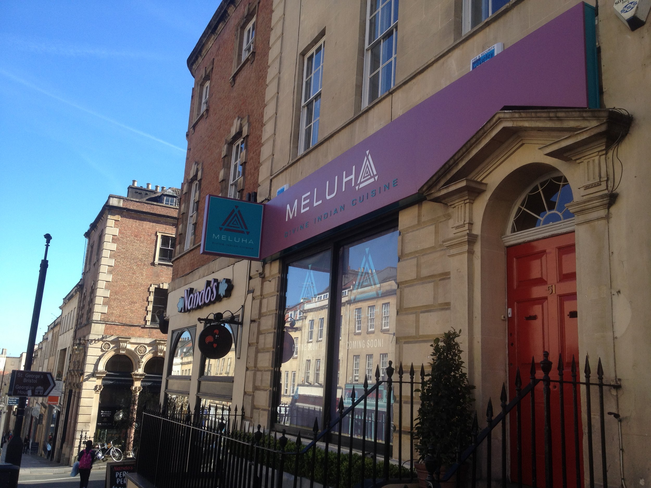 Street view of a building with a large purple sign for Meluha Indian Cuisine, a restaurant, and a red door. Other storefront signs are visible, including Nando's. Pedestrians and bicycles are on the sidewalk, with a clear blue sky overhead.