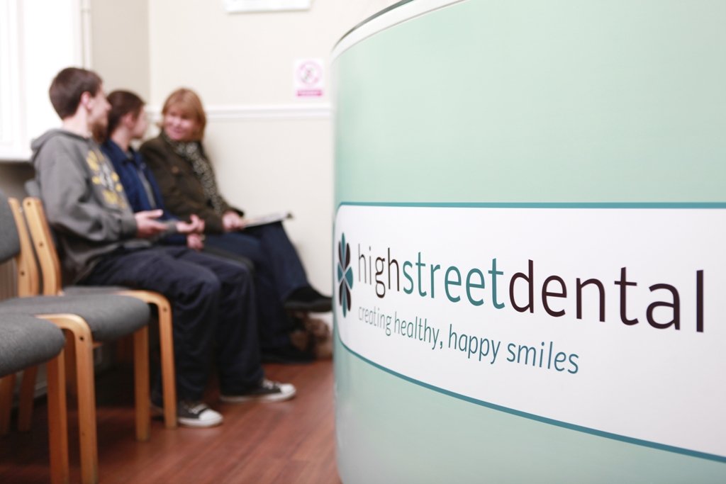 A reception area at High Street Dental with three people sitting on chairs in the waiting room, engaging in conversation. The sign in the foreground displays the dental office's name and slogan.